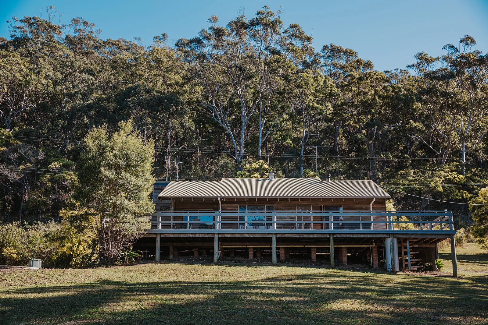 A wooden house on stilts with a metal roof and a spacious balcony, surrounded by a grassy area and dense trees in the background under a clear blue sky | Goolawah Retreat Crescent Head