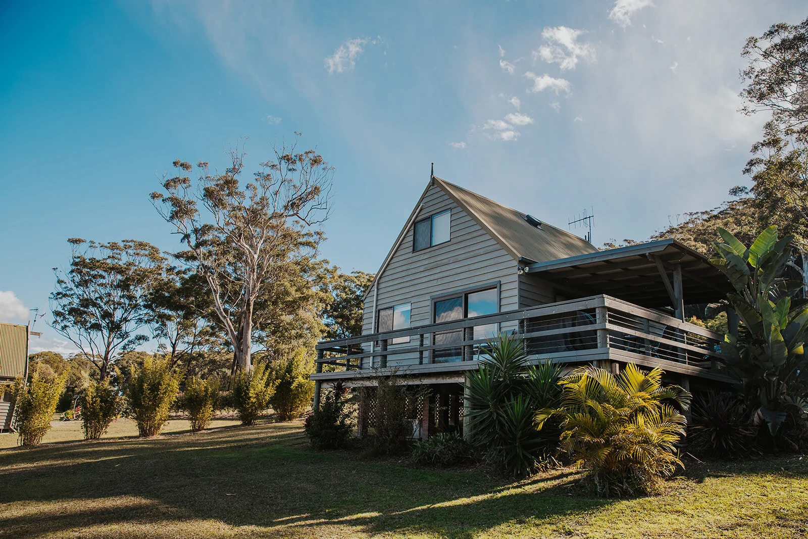 A wooden house with a deck, surrounded by trees and plants, under a blue sky with some clouds  | Goolawah Retreat Crescent Head