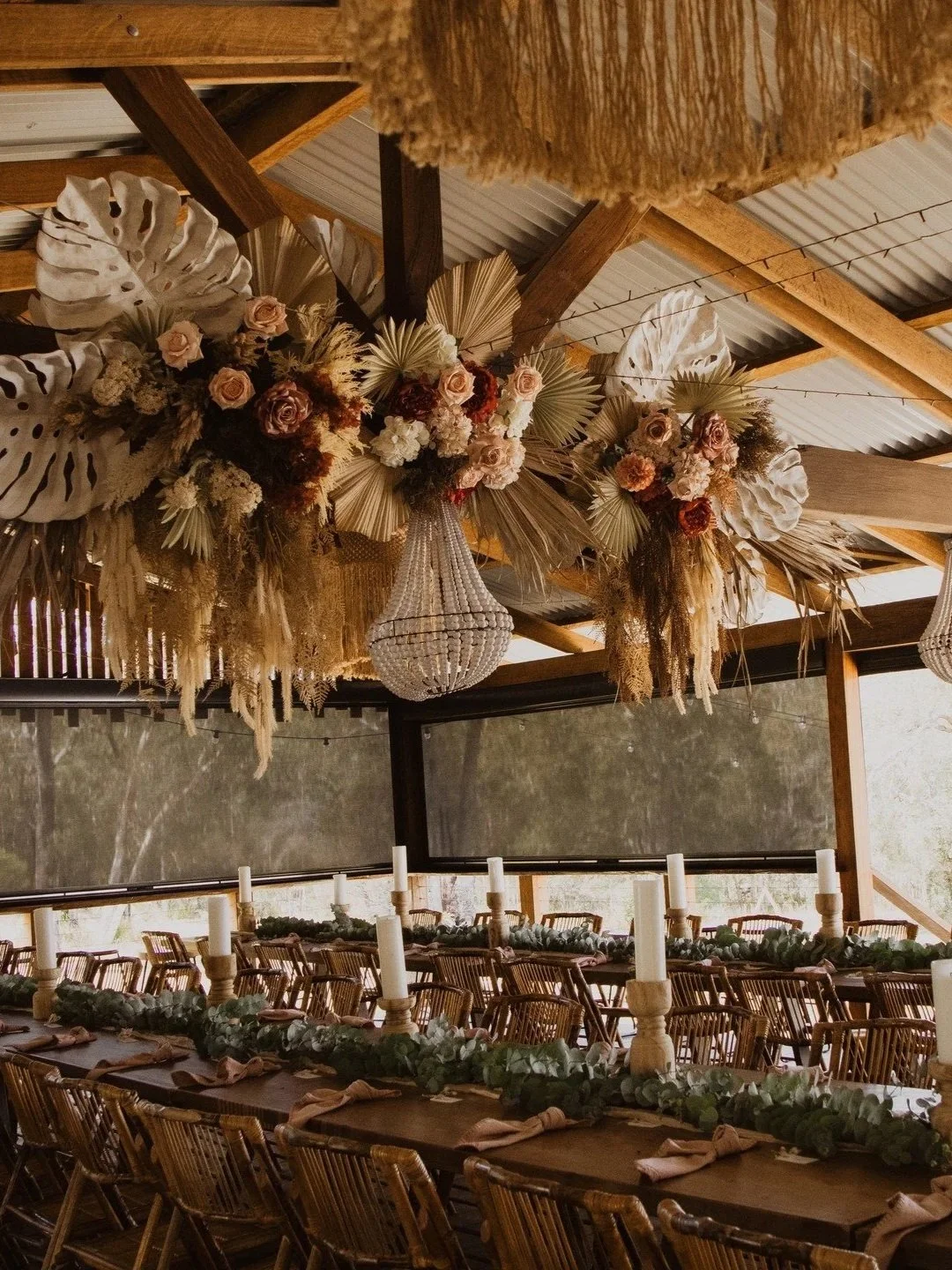 A decorated indoor event space with a floral and foliage ceiling arrangement. Long tables set for a gathering with candles and greenery, large windows with views of trees outside.