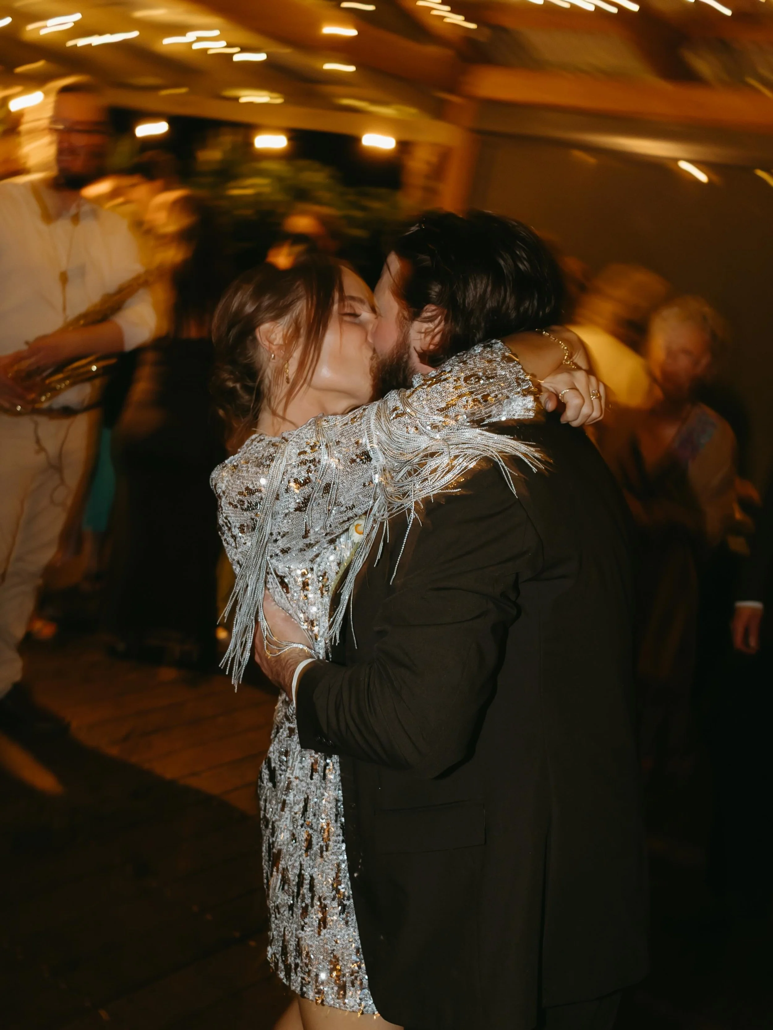 A couple kissing at a party or celebration, with other people in the background. The woman is wearing a sparkly, sequined dress with fringe details, and the man is dressed in a dark suit. The atmosphere appears lively with warm lighting and blurred m