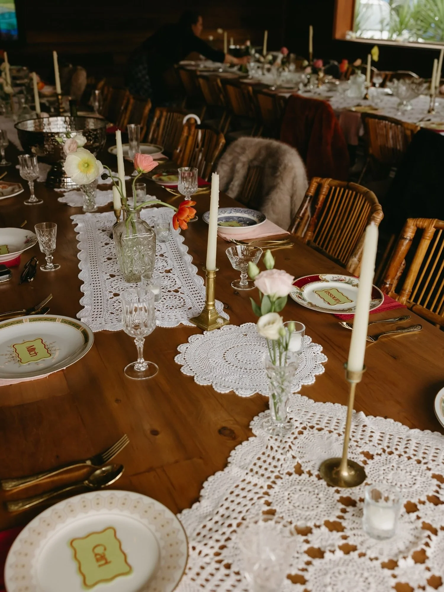 A dining table set for a meal with white lace table runners, floral centerpieces, candles, fine china, crystal glasses, and silverware. A person is arranging items in the background near a window with greenery outside.| Goolawah Retreat Wedding 