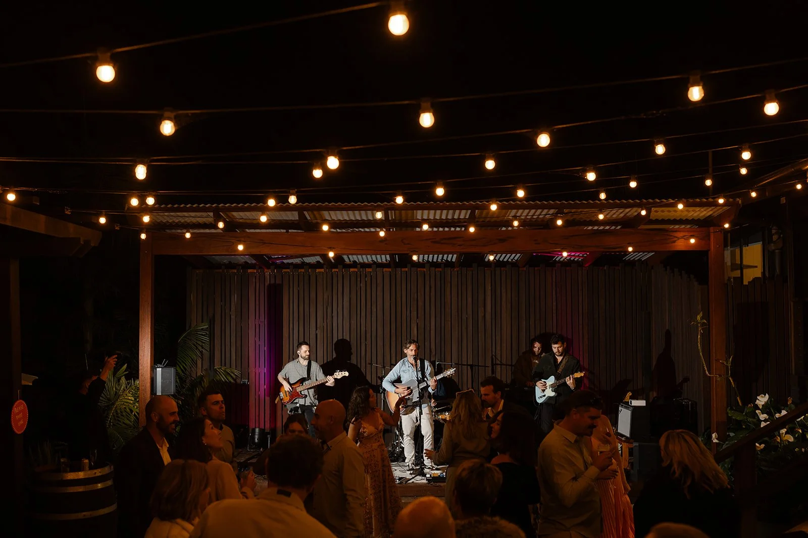 A band performing on a stage at an indoor venue with string lights overhead and an audience watching.