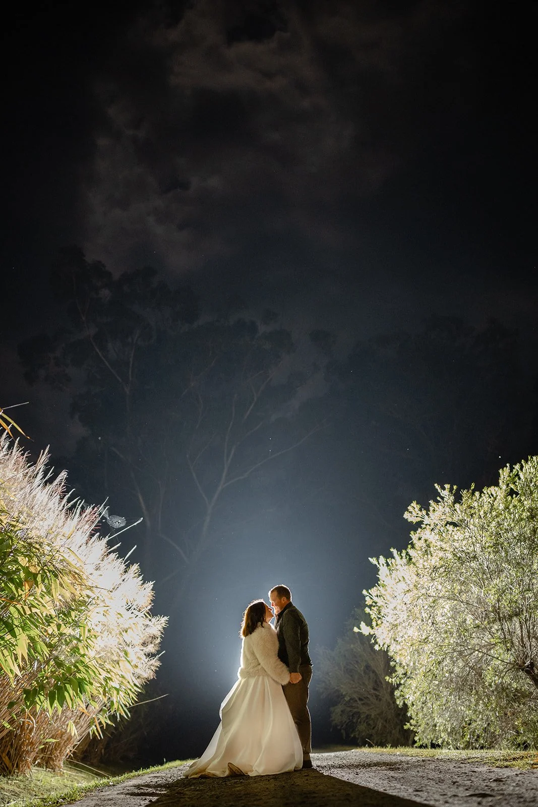 A newlywed couple stands close together at night, holding hands and sharing a moment beneath the moon and stars, surrounded by illuminated trees.| Goolawah Retreat Wedding Crescent Head