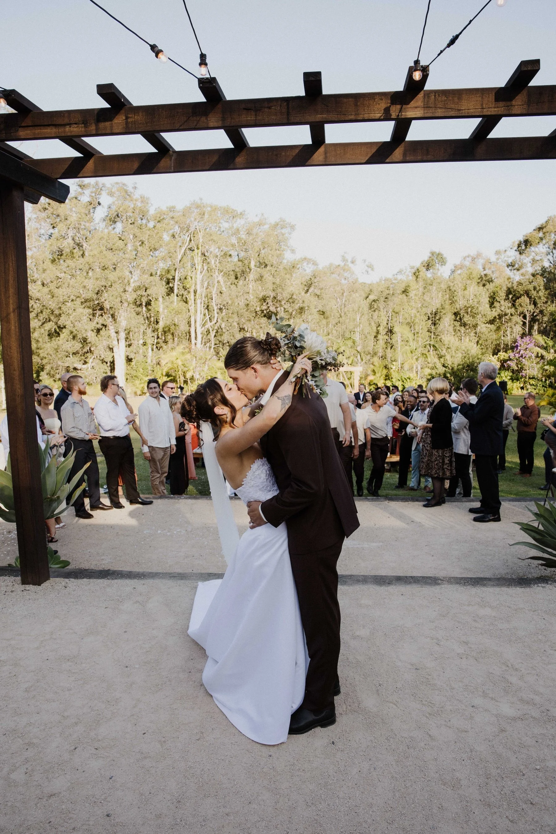 A bride and groom share a kiss during their outdoor wedding ceremony under a wooden arch, with guests watching and clapping in the background on a sunny day surrounded by trees.