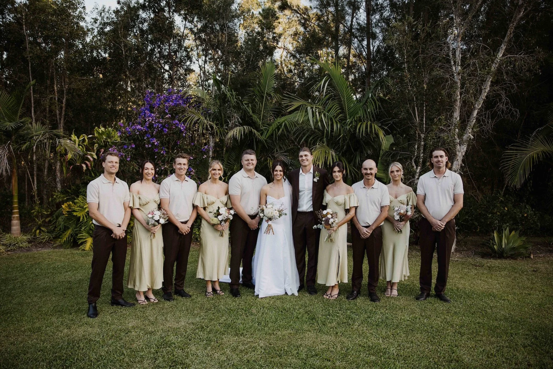 Group of people dressed in formal attire, including women in matching beige off-shoulder dresses holding bouquets of flowers, and men in light-colored shirts and dark trousers, standing outdoors in front of lush green plants and trees, likely at a wedding celebration.