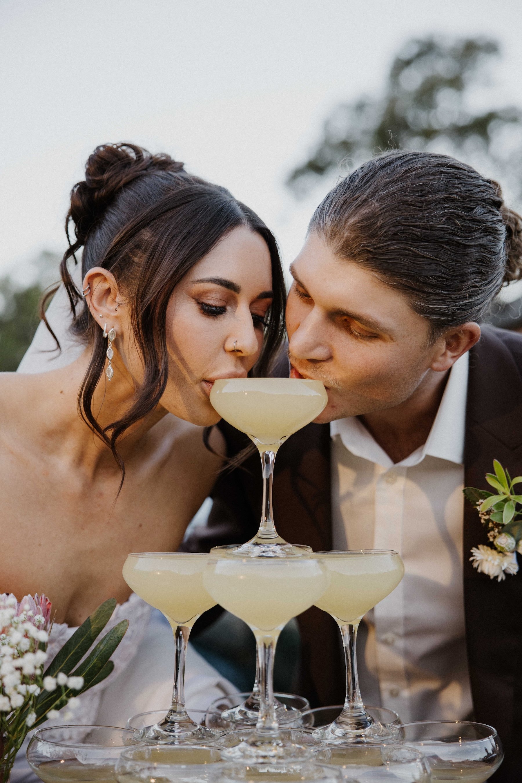 A couple with dark hair, dressed elegantly, is drinking champagne from a tiered champagne tower at a wedding celebration outdoors.