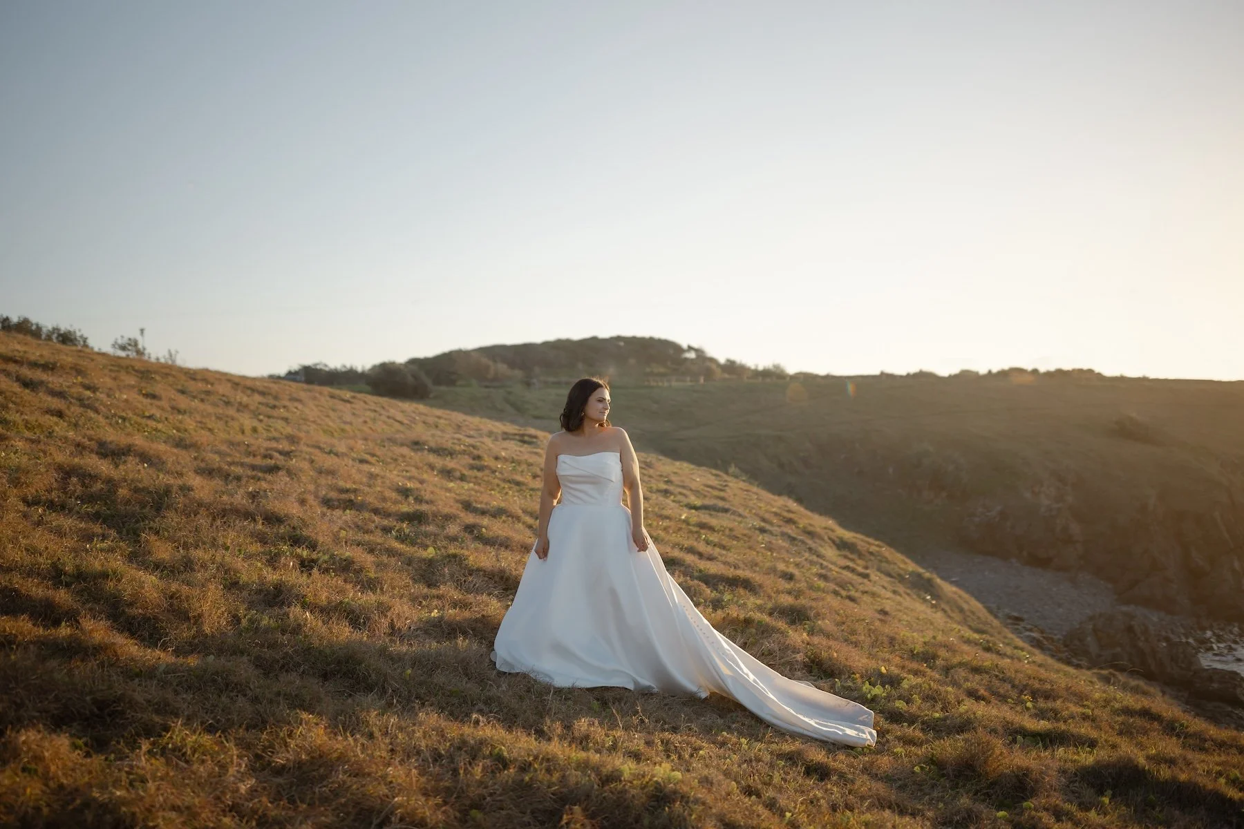 A woman in a white wedding dress standing on a hillside during sunset with rolling hills in the background.| Goolawah Retreat Wedding Crescent Head