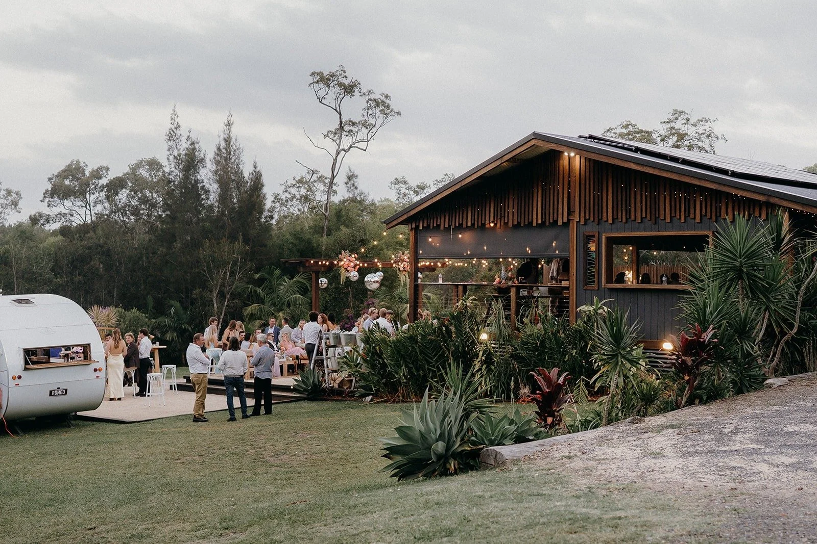 Outdoor gathering at a rustic modern venue with a wooden building, string lights, and a seated crowd, surrounded by lush greenery and trees.