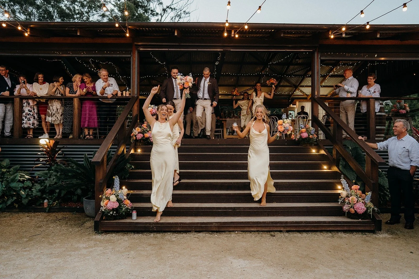Two women in cream-colored dresses celebrate on the steps of a wooden deck at a wedding reception, holding bouquets and glasses, with guests watching from above at dusk.