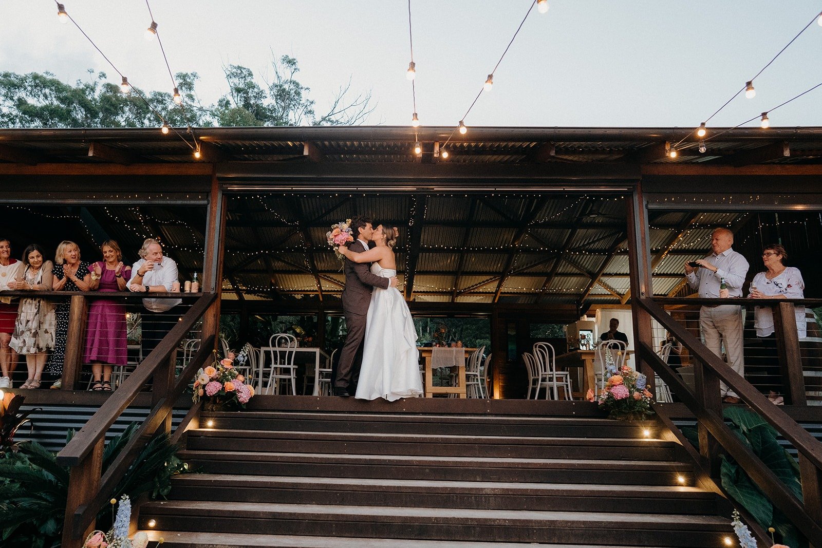 A bride and groom sharing a kiss during their wedding reception on an outdoor venue with guests watching. String lights hang above, and the setting includes floral arrangements and tables.