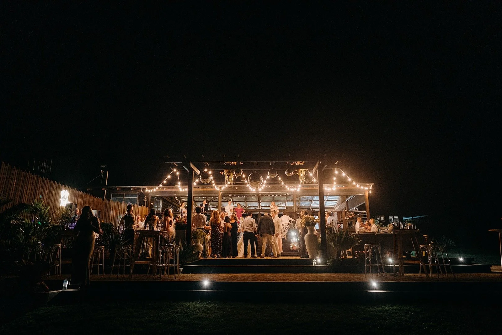 Nighttime outdoor party with people dancing and socializing under string lights at a decorated venue.