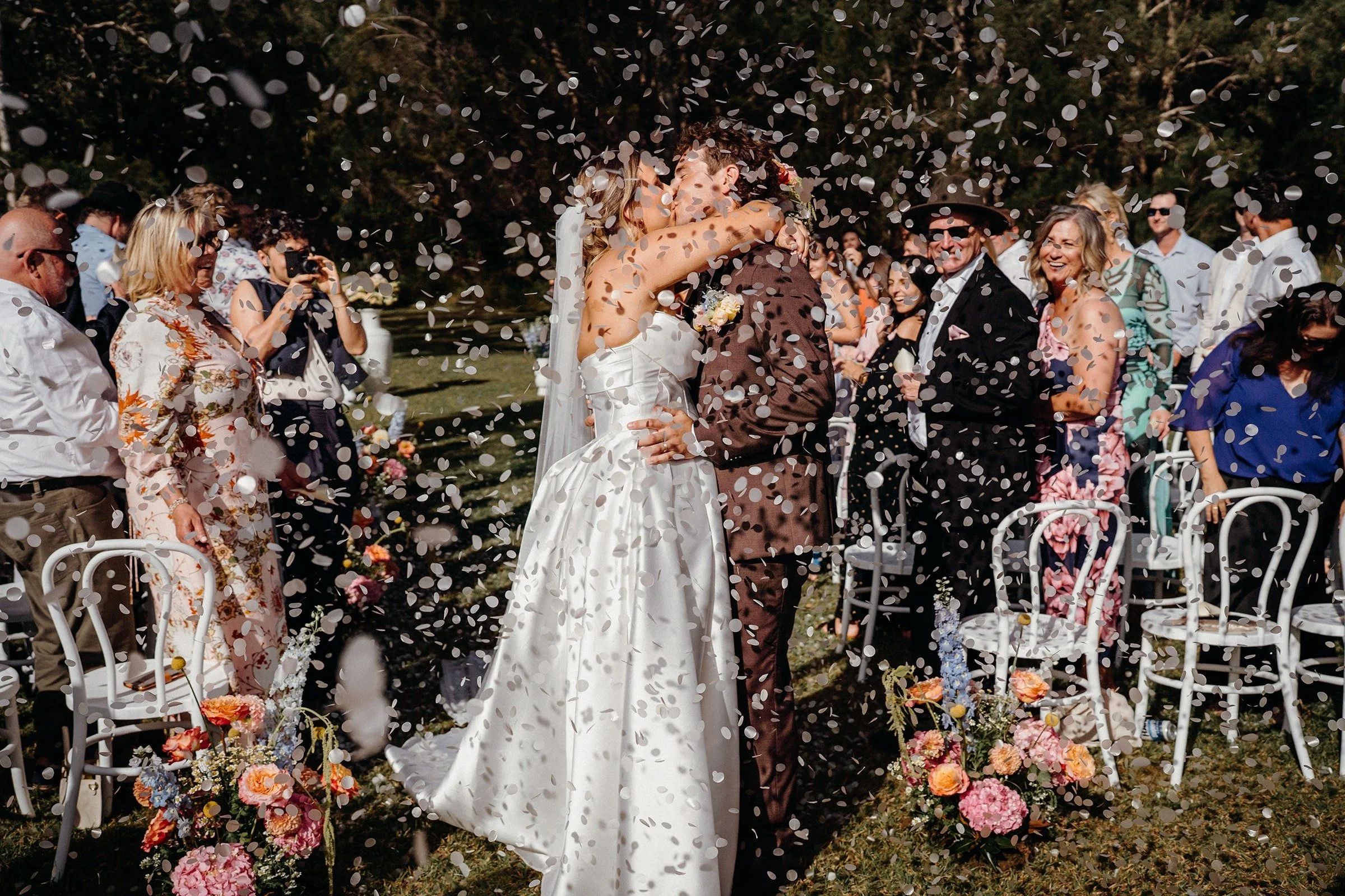A bride and groom share a kiss at their outdoor wedding ceremony amidst falling confetti, surrounded by seated guests, floral decorations, and lush greenery.| Goolawah Retreat Wedding Crescent Head