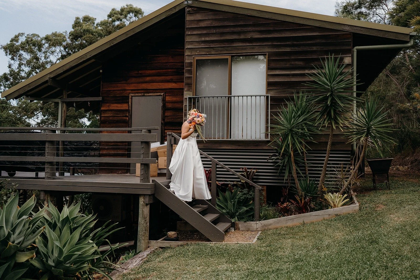 A woman in a wedding dress holding a bouquet walks down the stairs of a wooden house with a small balcony and surrounded by green plants.
