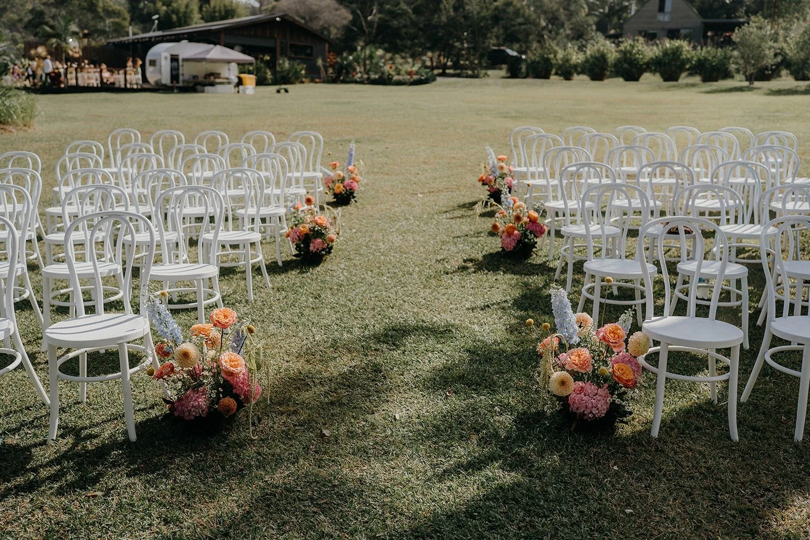 Outdoor wedding ceremony setup with white chairs arranged along a grass aisle, decorated with colorful flower arrangements.
