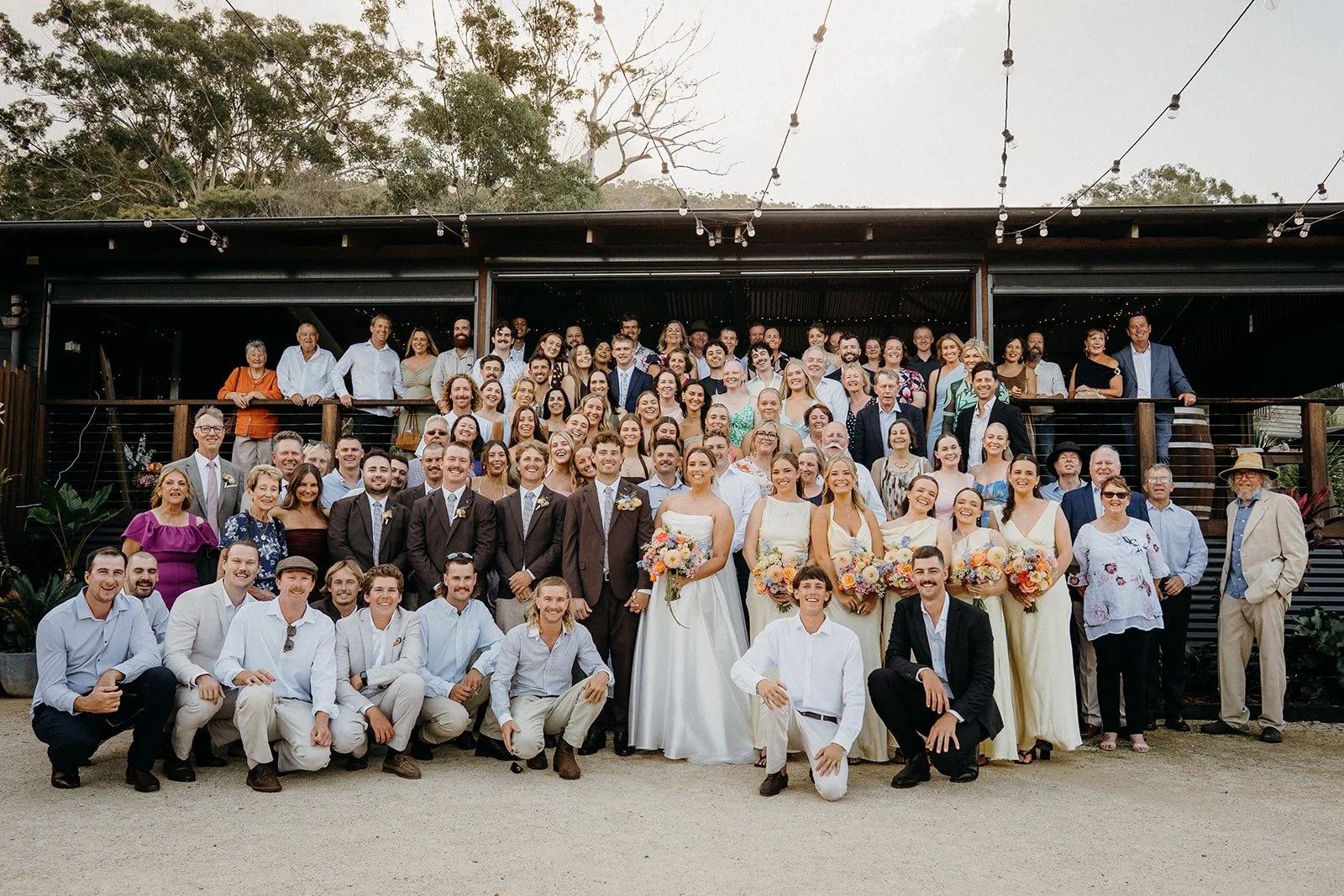 A large group of people at a wedding, with the bride and groom in front, surrounded by family and friends, outdoors in a natural setting.