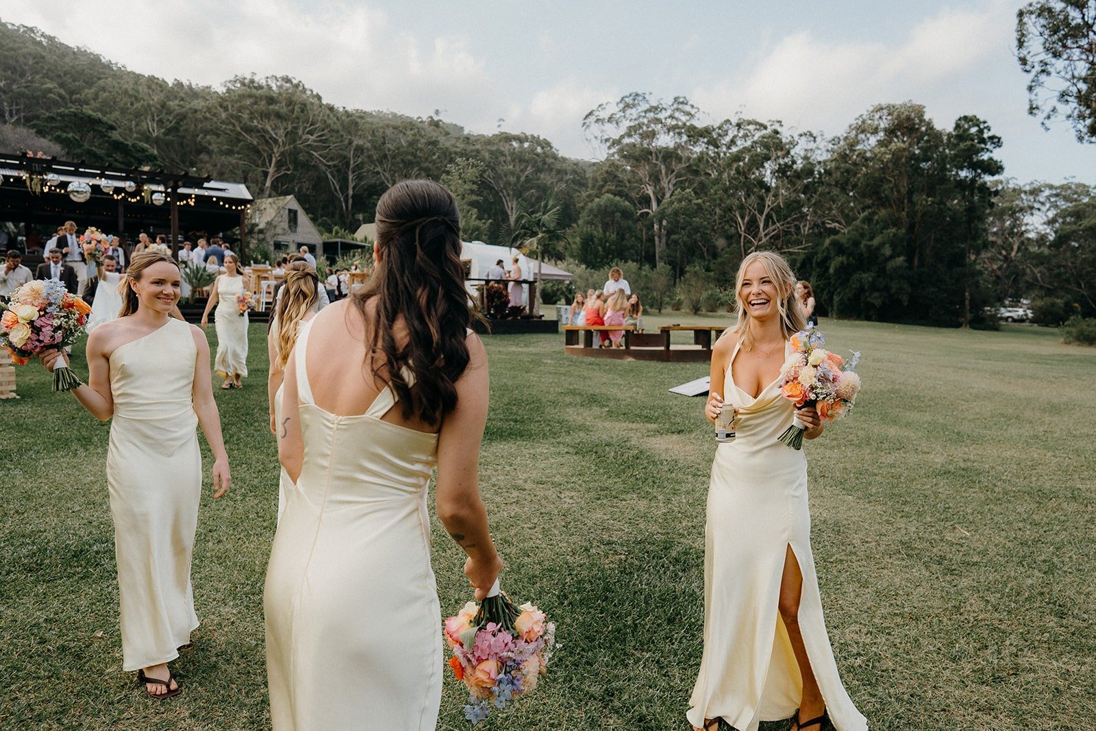 Three women in cream-colored dresses at an outdoor wedding reception, with one woman holding a bouquet and smiling at another woman who is also holding a bouquet, while guests and a band are in the background on a grassy field surrounded by trees.