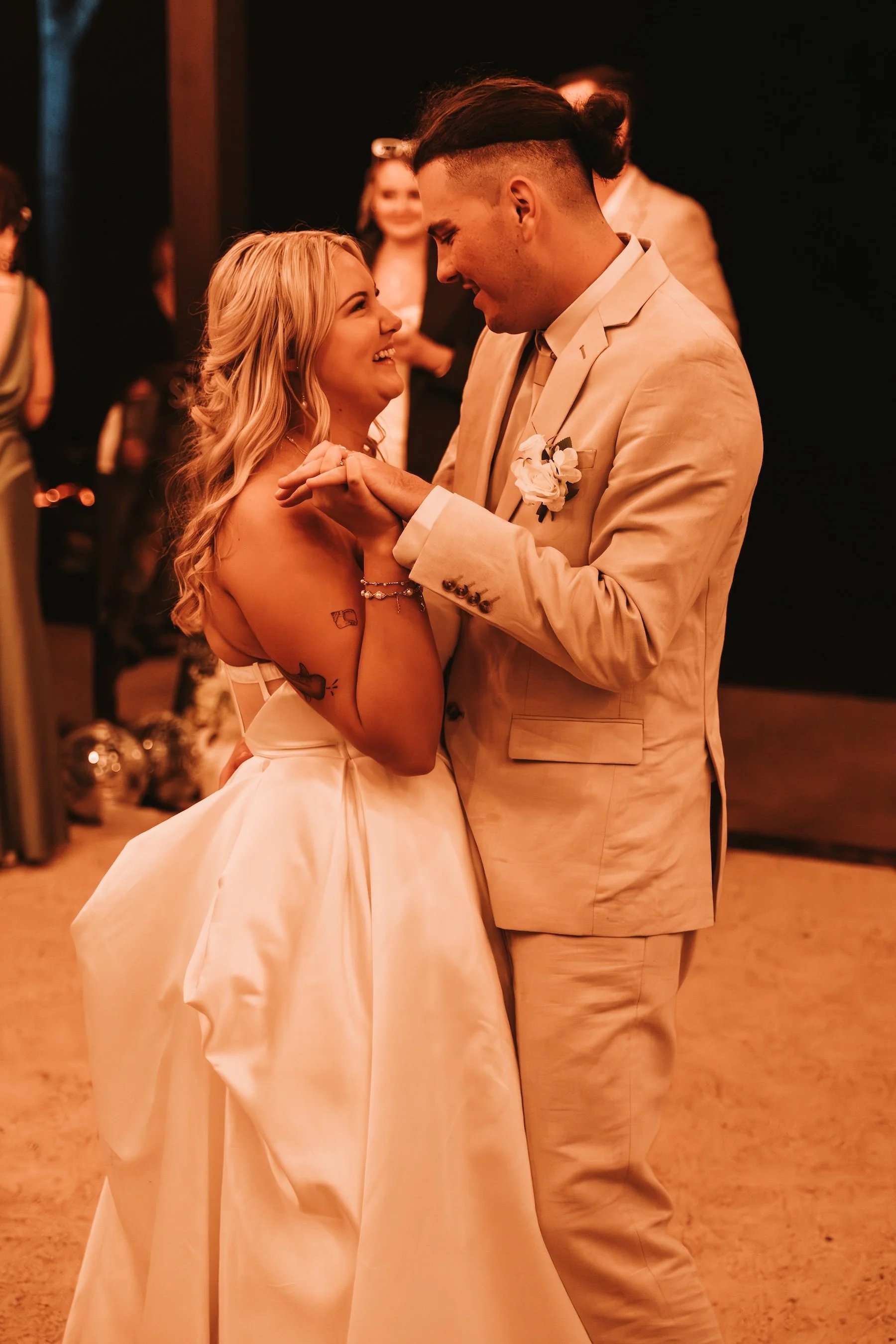 A couple dancing at a formal event, smiling at each other, with women and decorated tables in the background.
