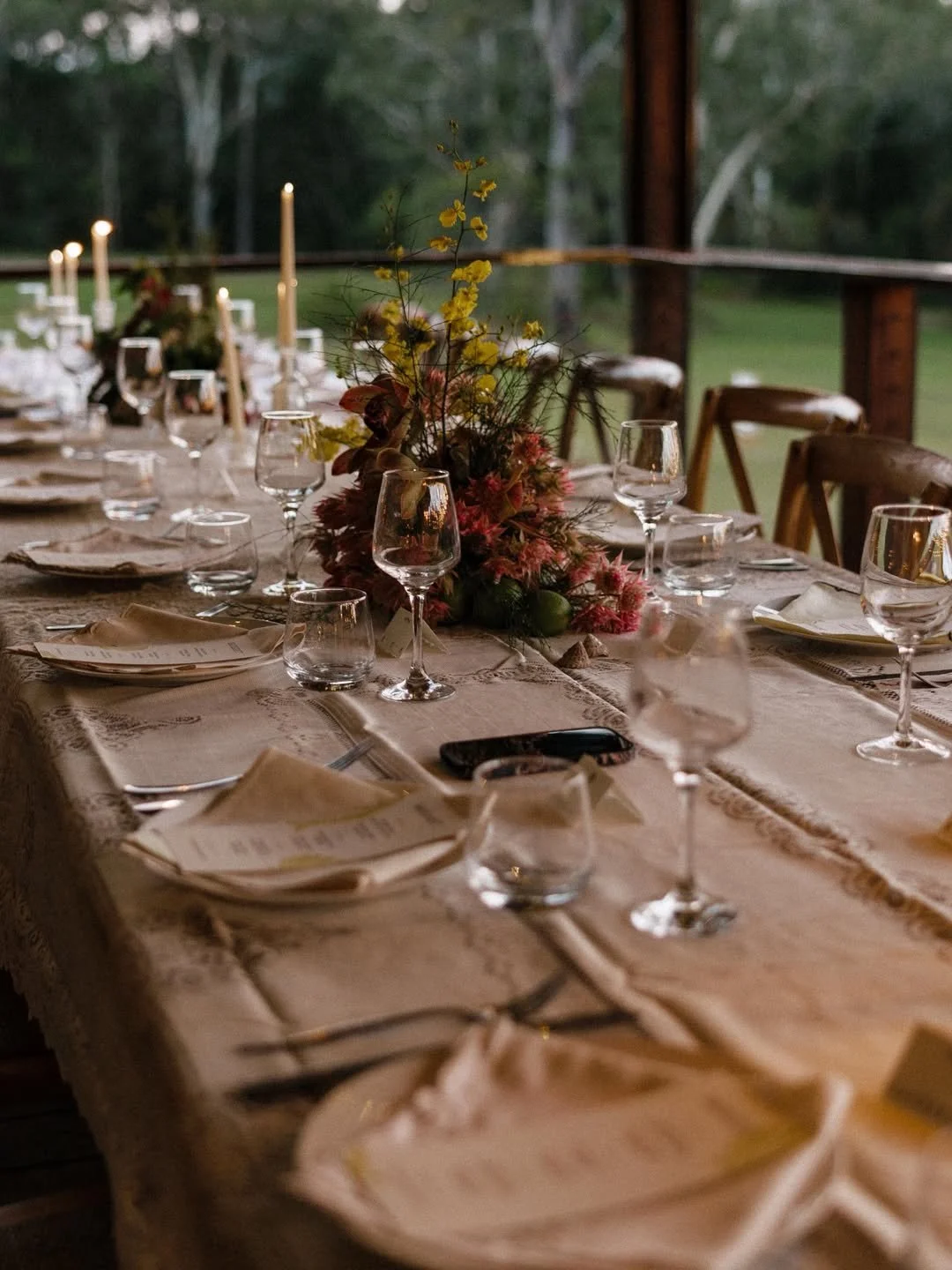 An elegantly set dinner table with multiple wine glasses, plates with folded napkins, and a large floral centerpiece, outdoors with greenery visible in the background.