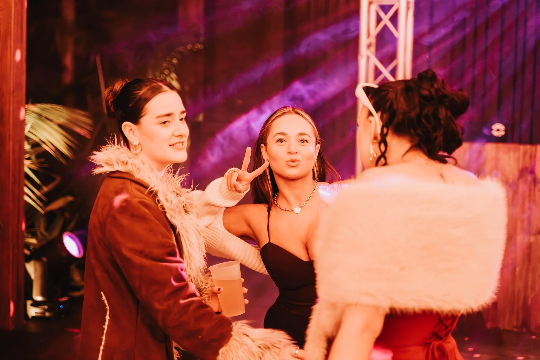 Three young women at a party or nightclub, taking a selfie together with colorful lighting in the background. One woman is making a peace sign and puckering her lips, while another holds a drink. They are dressed stylishly with accessories and fur-tr