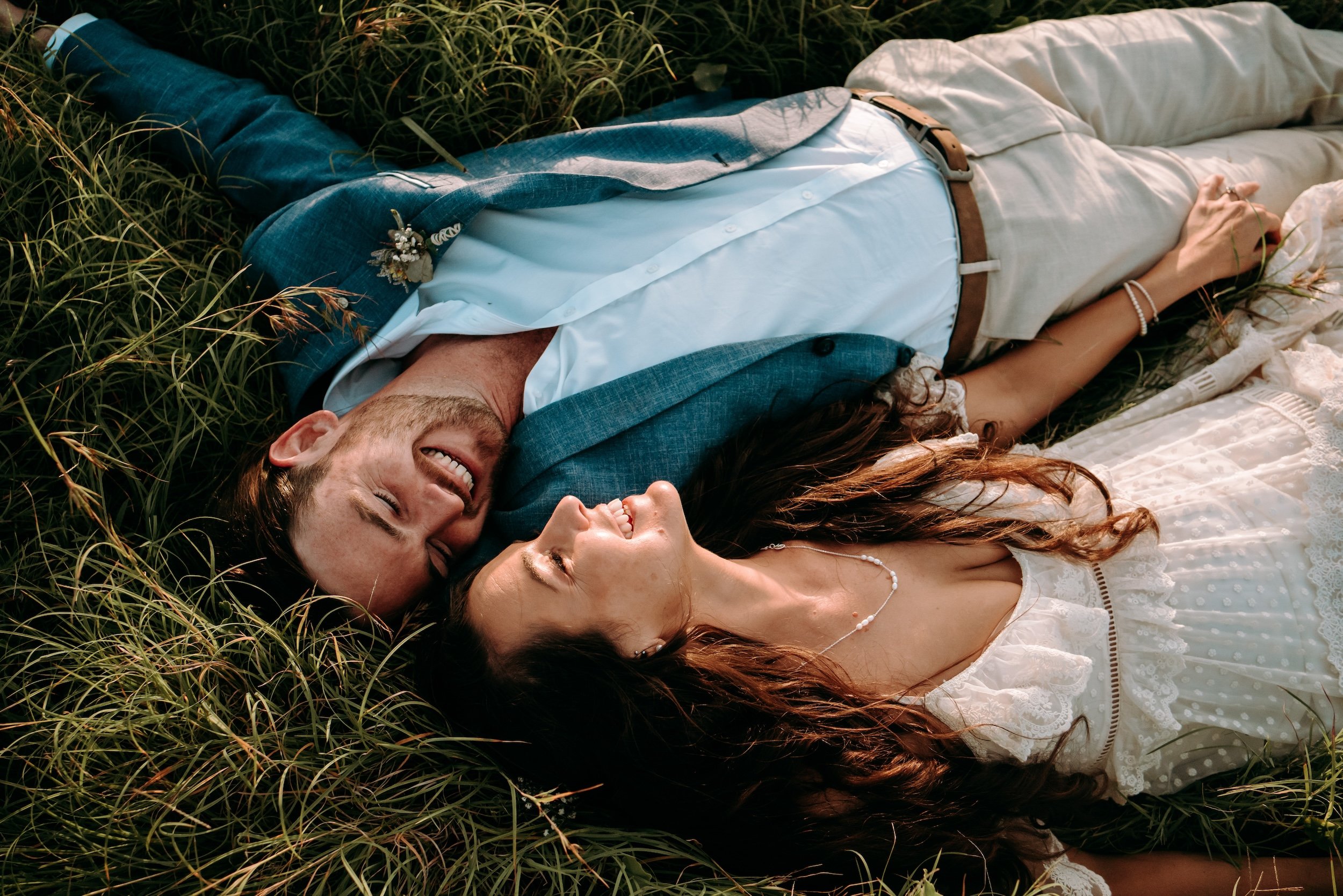 A couple lying outdoors in grass, smiling and looking at each other. The man is wearing a blue blazer, white shirt, and jeans. The woman is in a white dress with lace detail, wearing a pearl necklace and bracelet.
