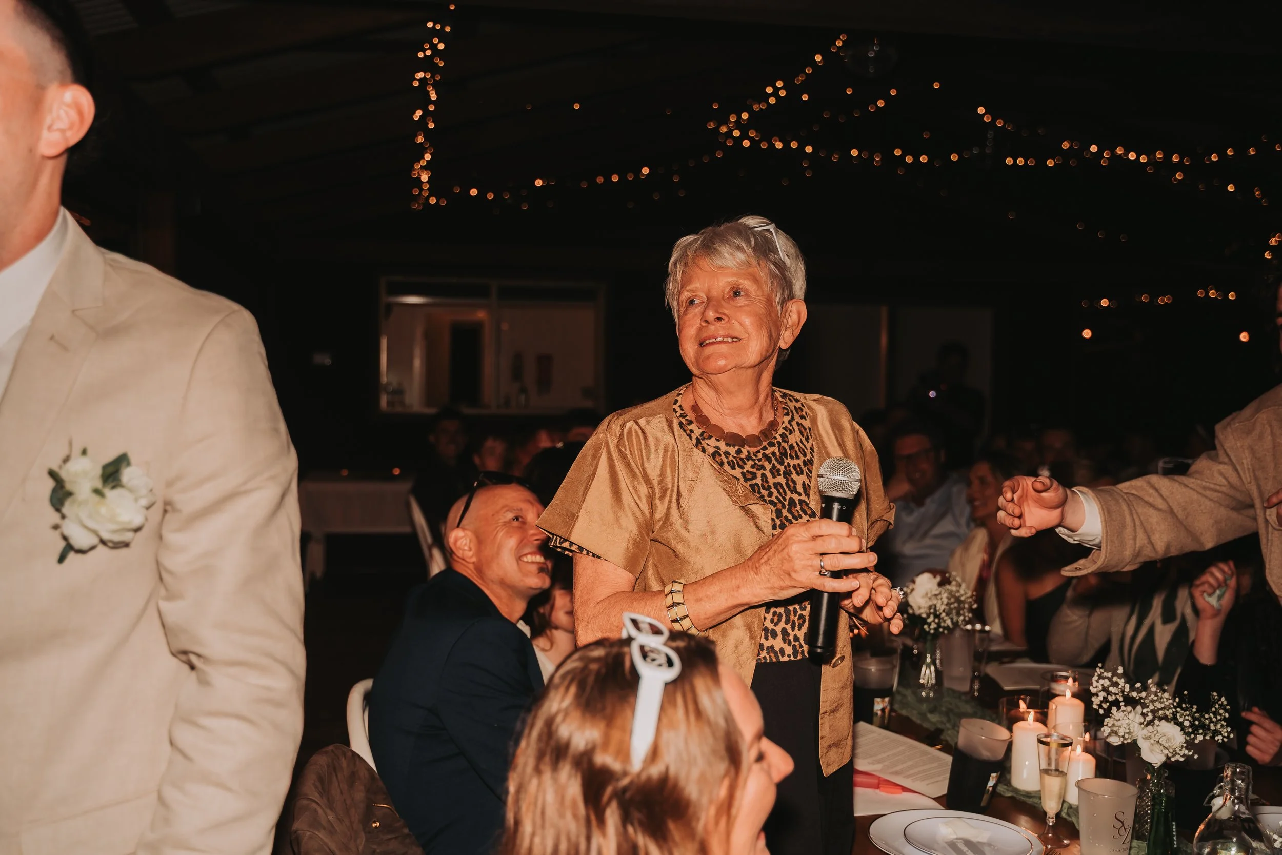 An elderly woman with short gray hair and wearing a leopard print top with a beige jacket, is speaking into a microphone at a dinner or celebration event. She is holding the microphone with both hands, and there are people seated around a table with 