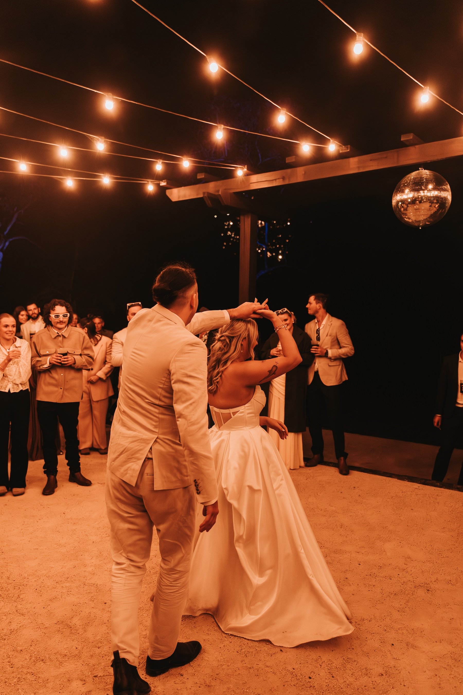 People dancing at a wedding reception outdoors at night under string lights and a disco ball.