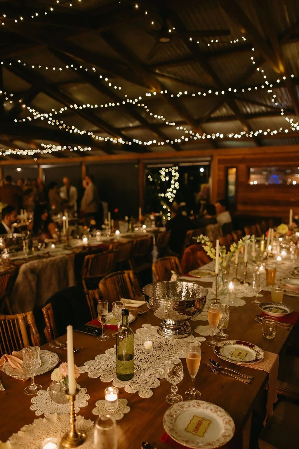 A warmly lit, decorated dining area with string lights on the ceiling, set for a banquet with candles, wine bottles, plates, glasses, and floral centerpieces.