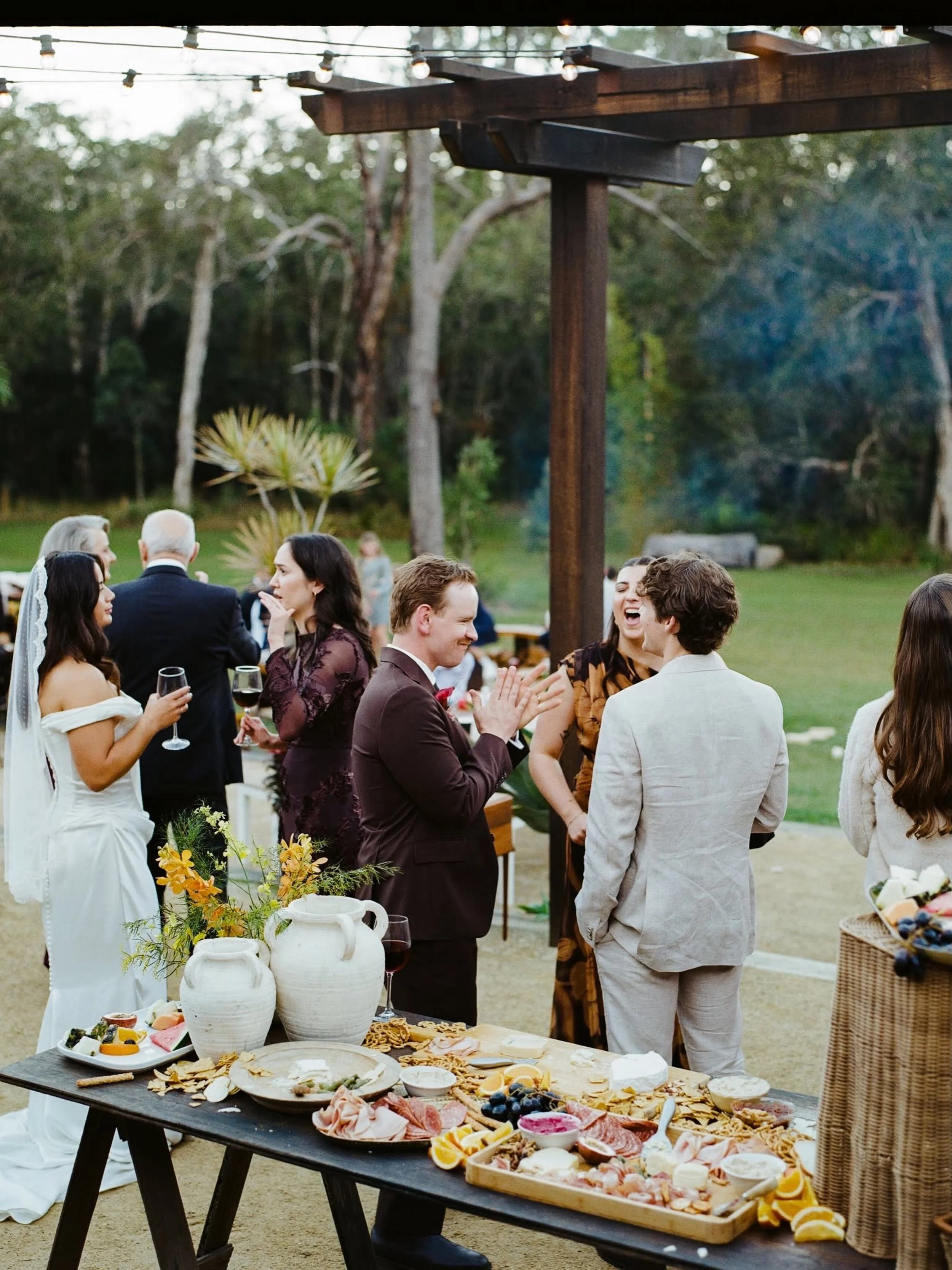 Guests dressed in formal attire at an outdoor wedding reception, mingling and celebrating under string lights with a lush green forest background. A table with cheeses, fruits, and crackers is visible in the foreground.