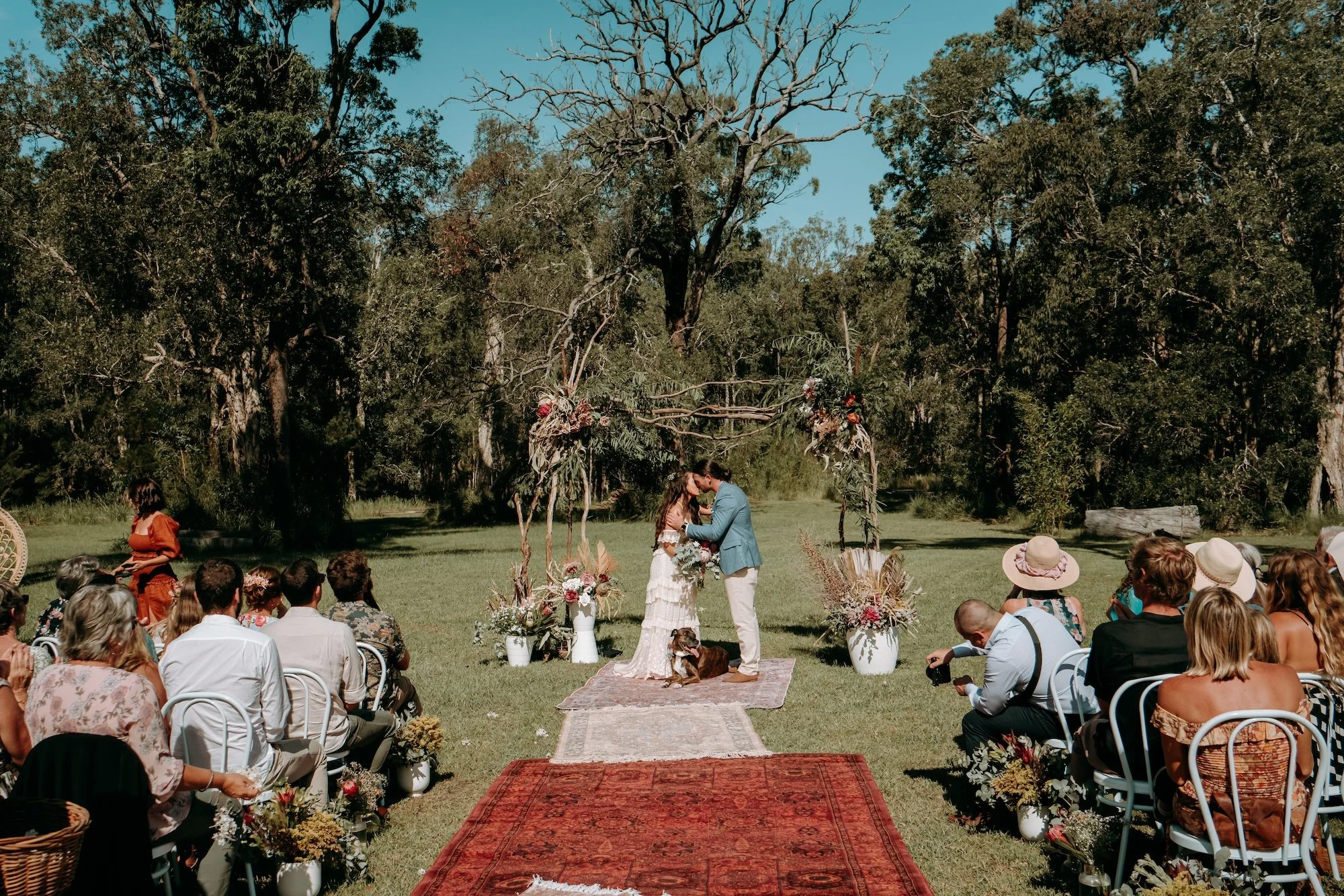 An outdoor wedding ceremony in a lush green setting with trees and blue sky. The couple stands before an officiant, surrounded by floral arrangements, with guests seated on either side. The bride and groom are kissing, and the scene includes guests w