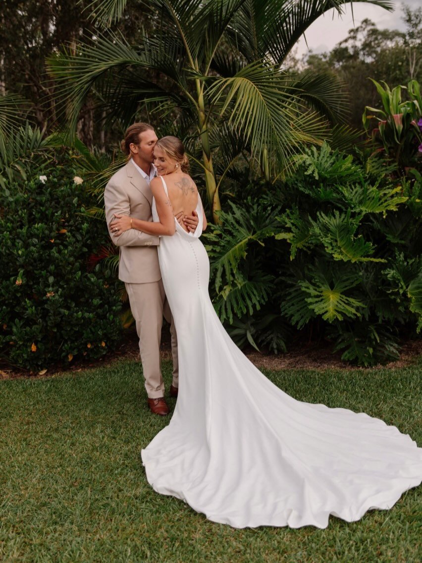 A bride and groom embrace each other in a lush outdoor setting with tropical plants, the bride in a white wedding gown with a long train, and the groom in a beige suit.| Goolawah Retreat Wedding Crescent Head