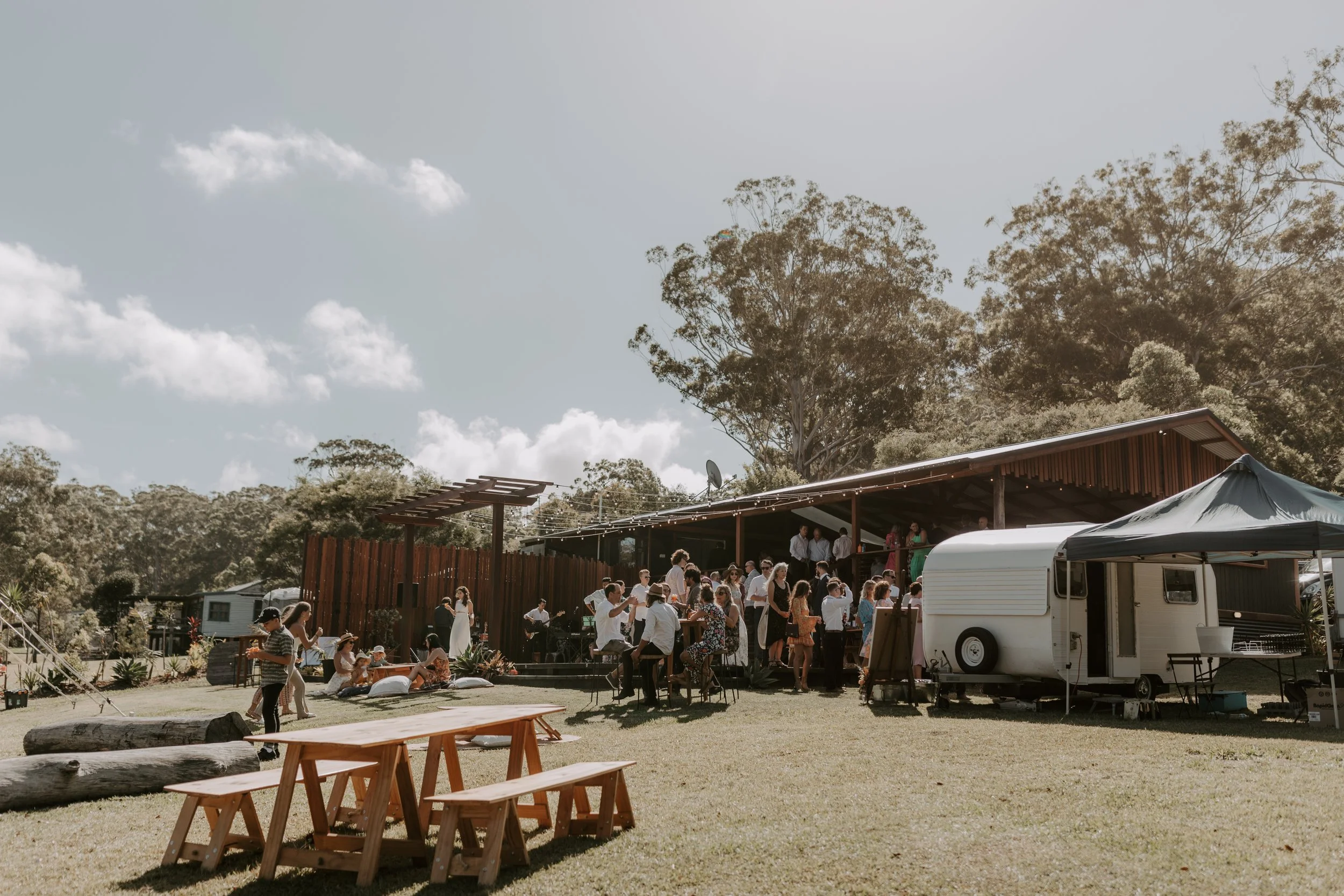 Outdoor gathering with people at a celebration or event near a wooden building with trees and a partly cloudy sky in the background. | Goolawah Retreat Crescent Head