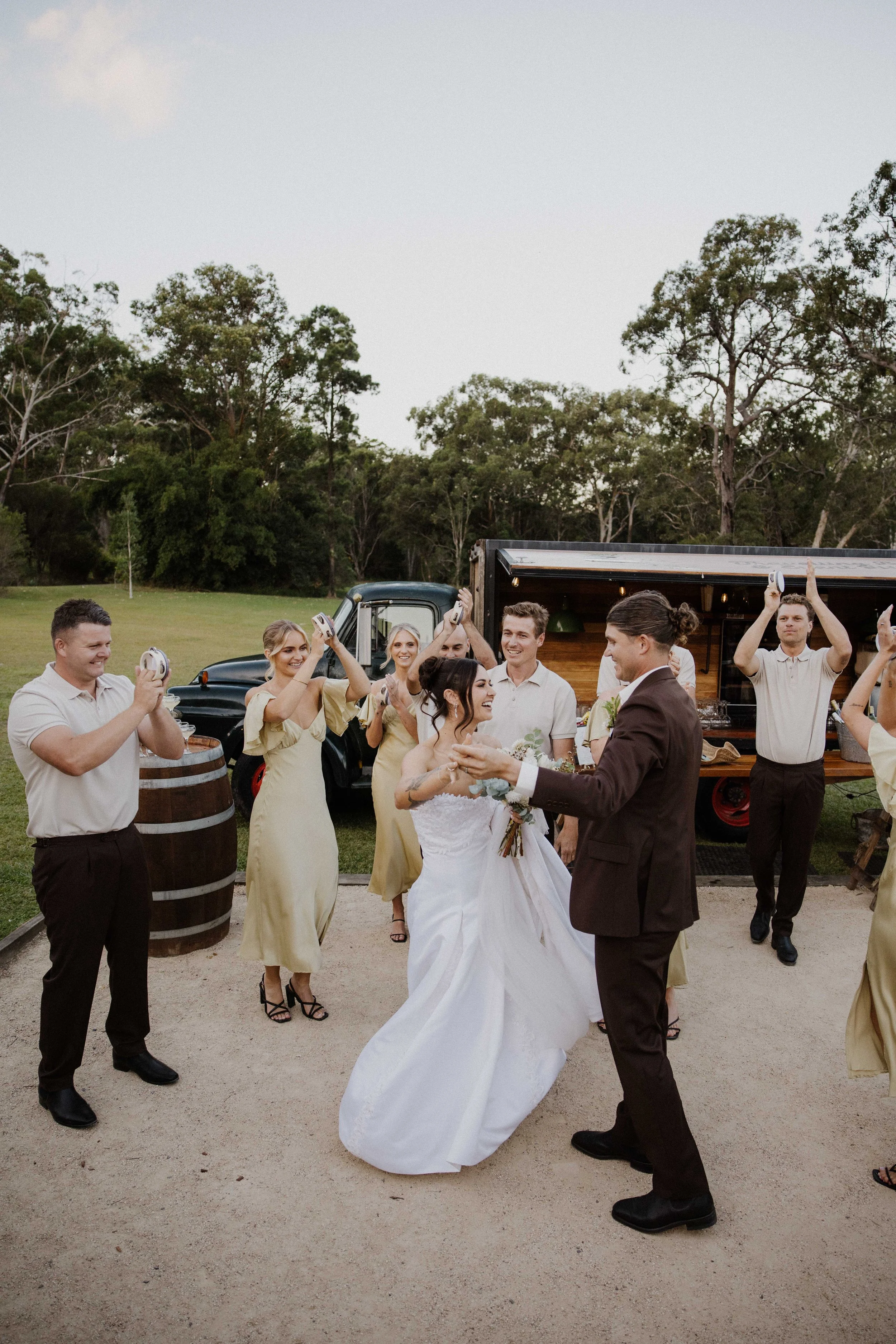 A wedding celebration outdoors with a bride and groom dancing among guests, some holding plates or glasses, and a vintage truck serving as a backdrop, surrounded by trees.