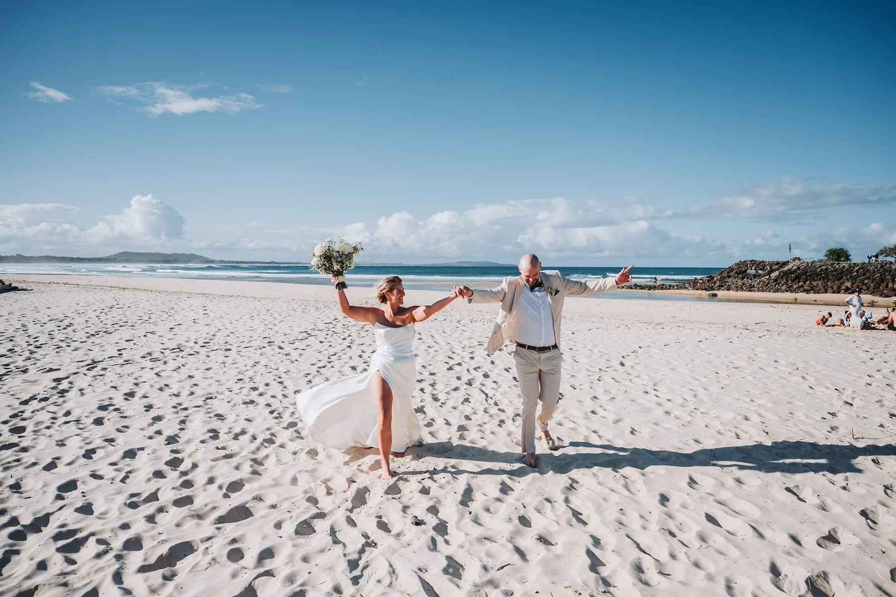 A newlywed couple in wedding attire dancing on a sandy beach. The bride is holding a bouquet and wearing a strapless white dress with a high slit, while the groom is in a light-colored suit with a bow tie. They are joyfully holding hands and smiling. The ocean and partly cloudy sky are in the background, with a few people sitting on the sand and rocks along the shore.