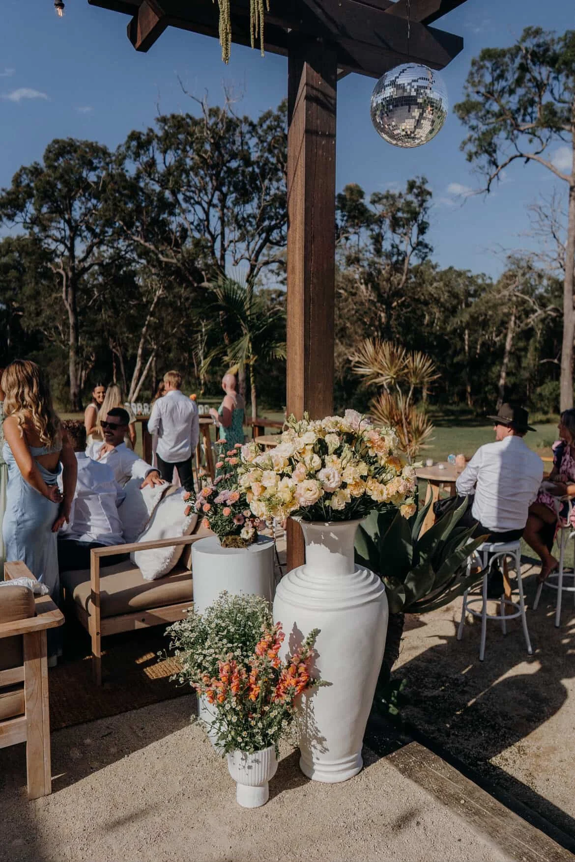 People gathered outdoors for a celebration, with large white floral arrangements in vases in the foreground, and a disco ball hanging from a wooden structure.