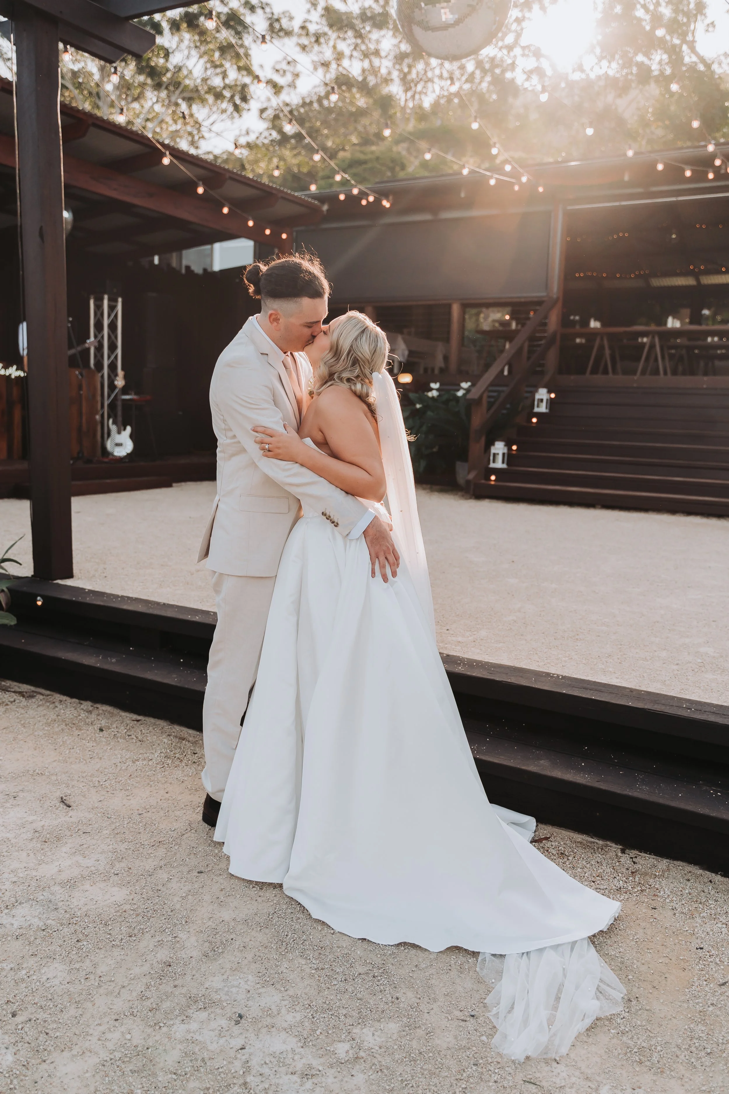 A couple in wedding attire sharing a kiss outdoors at sunset, with string lights and a stage in the background.