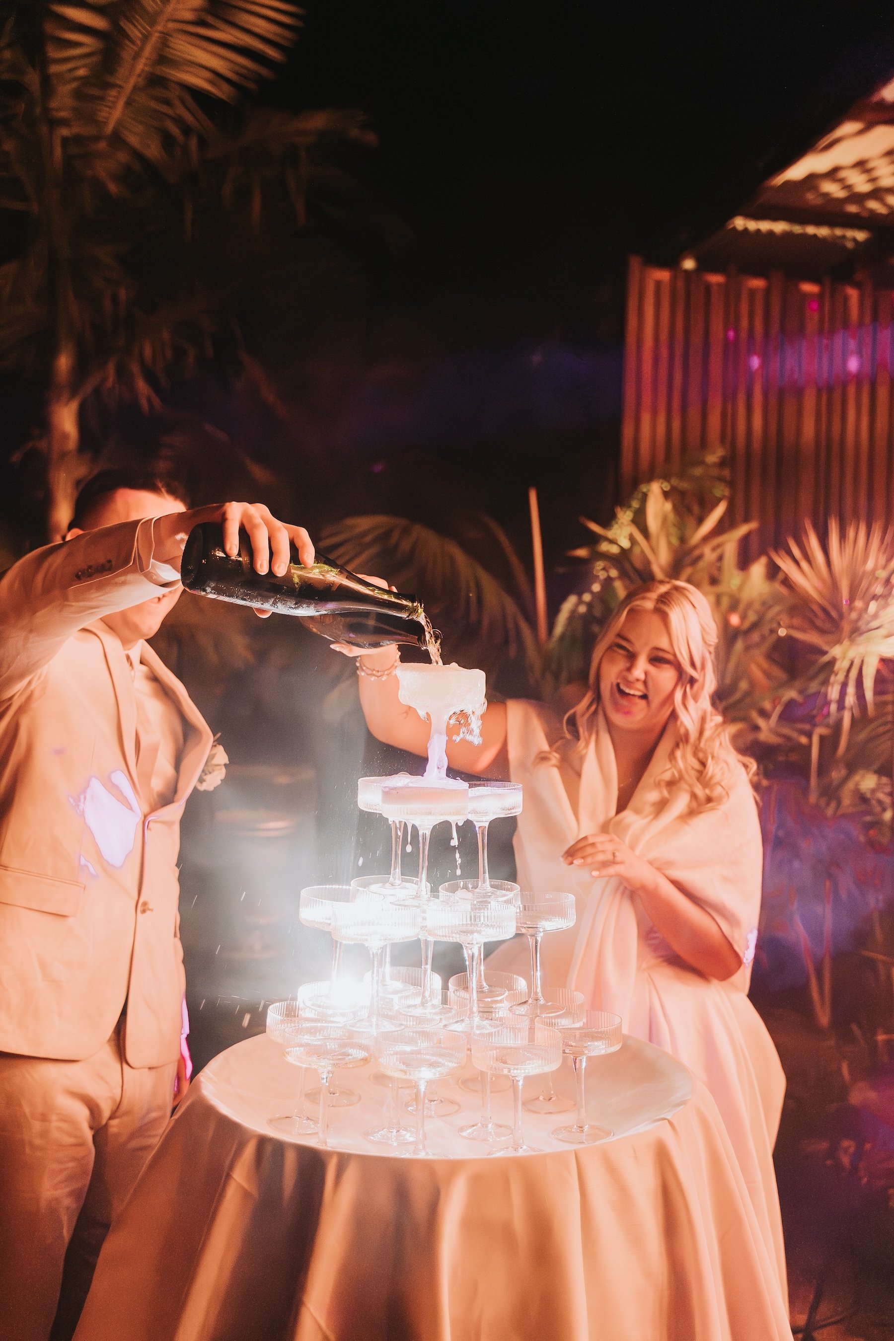 People celebrating at a party with a champagne tower, a woman pouring champagne and another woman smiling, in a decorated outdoor setting with palm plants and ambient lighting.