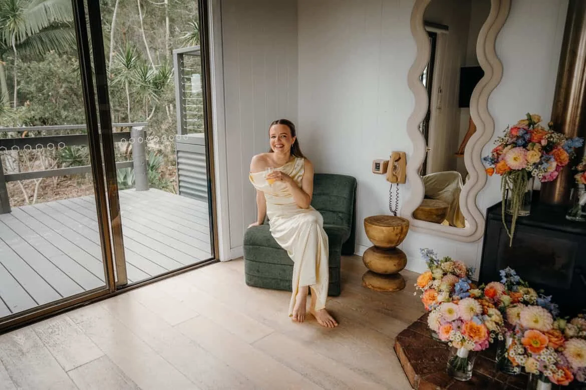 A woman in a cream-colored dress sitting on a green chair in a room with a glass door leading to a patio with plants outside. The room has a large mirror, a telephone on the wall, a vase with colorful flowers, and a wooden side table.