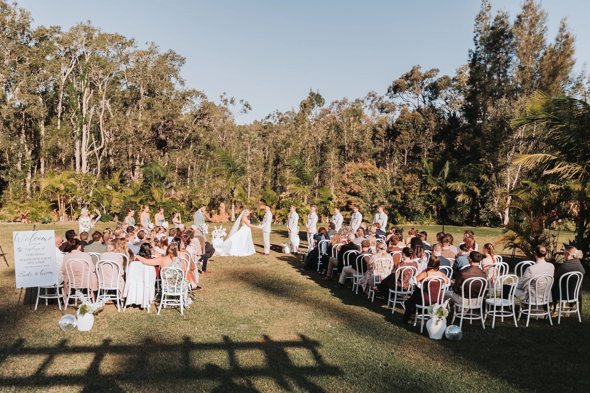 An outdoor wedding ceremony taking place in a lush green park with tall trees and clear sky in the background. The bride and groom are standing at the altar, exchanging vows. Guests seated on white chairs are watching the ceremony, with some wearing 