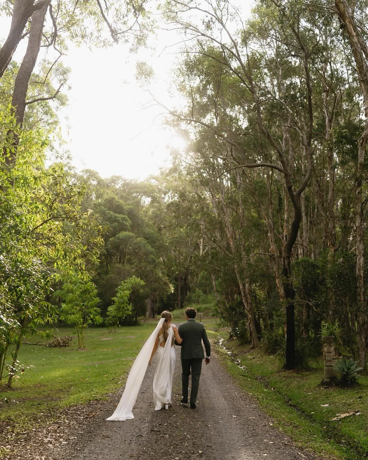 A bride and groom walking hand in hand along a wooded path during sunset, surrounded by lush green trees and foliage.