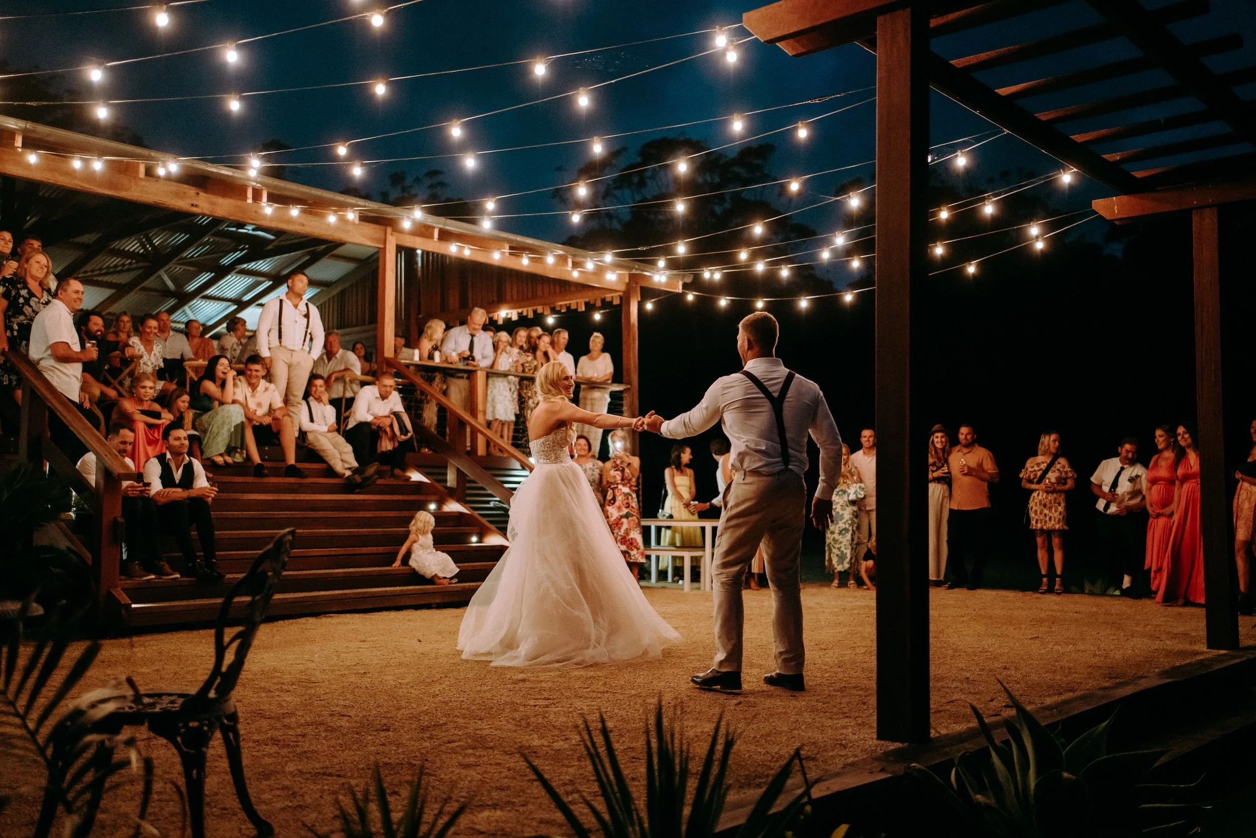 A nighttime outdoor wedding reception with string lights, a bride and groom dancing, and guests watching them, surrounded by a wooden patio structure and dark sky. | Goolawah Retreat Crescent Head