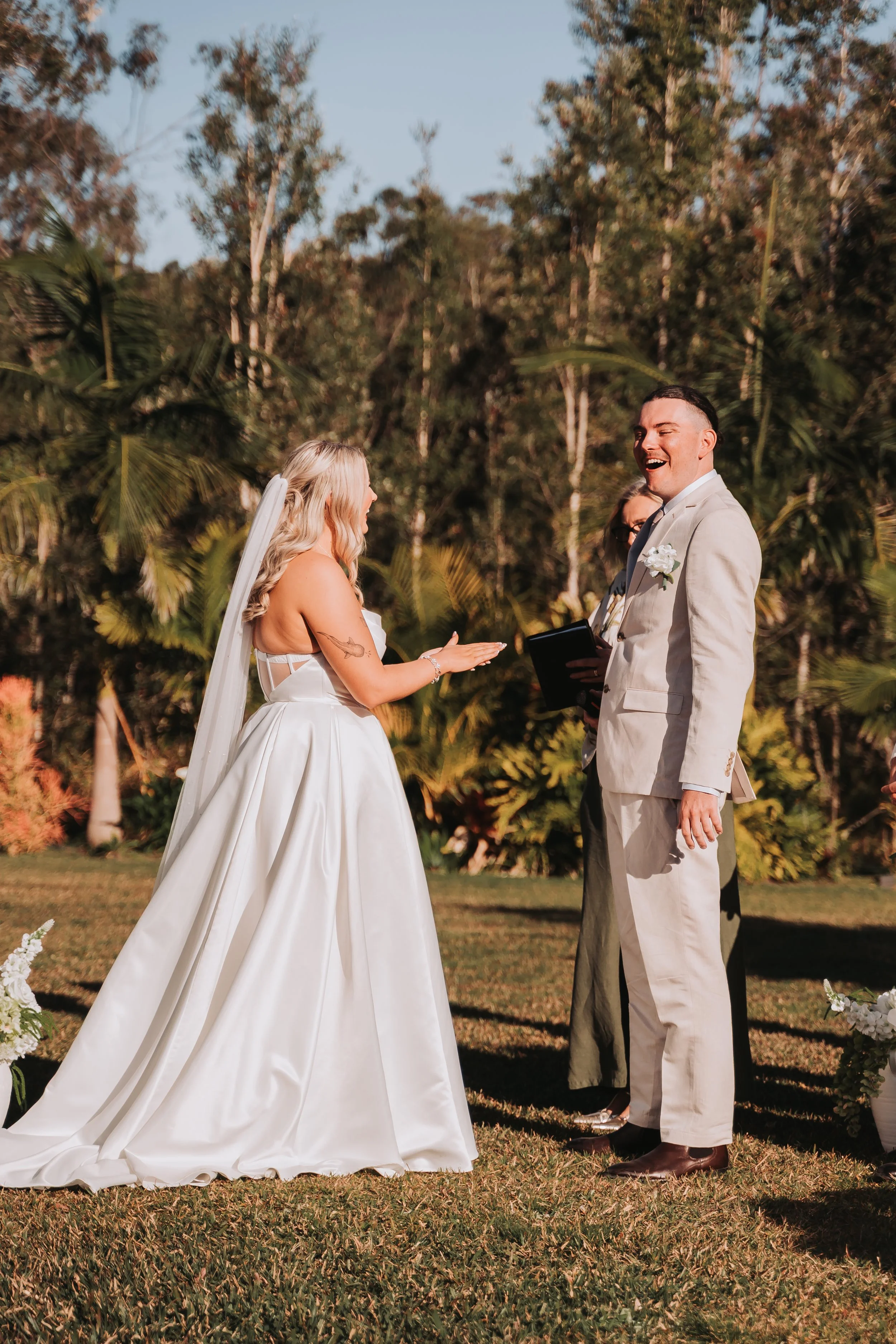 A wedding ceremony outdoors with a bride and groom exchanging vows, surrounded by trees and grass, during sunset.