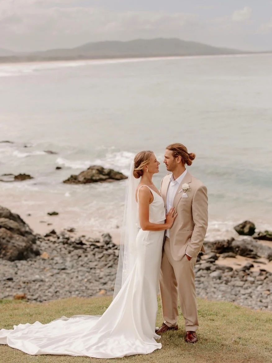 A bride and groom standing on a grassy area by the beach, facing each other, with the bride in a white wedding dress and the groom in a beige suit, holding hands, and looking into each other's eyes. | Goolawah Retreat Wedding Crescent Head