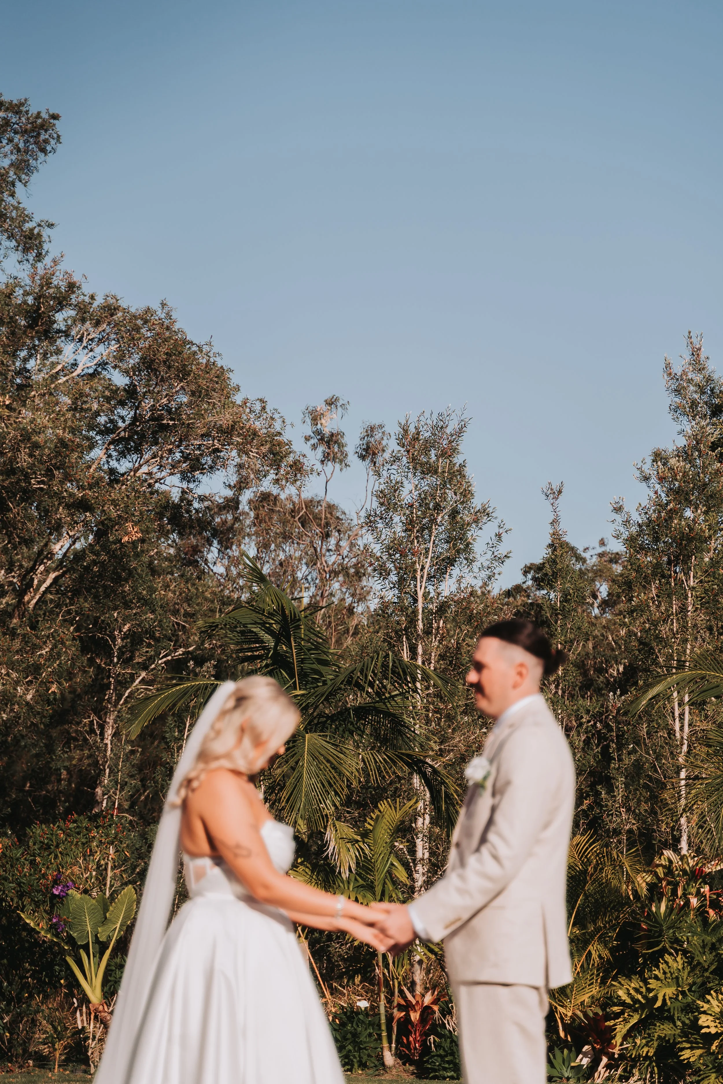 A wedding ceremony outdoors with a bride and groom holding hands and facing each other, surrounded by lush greenery and trees, under a clear blue sky.