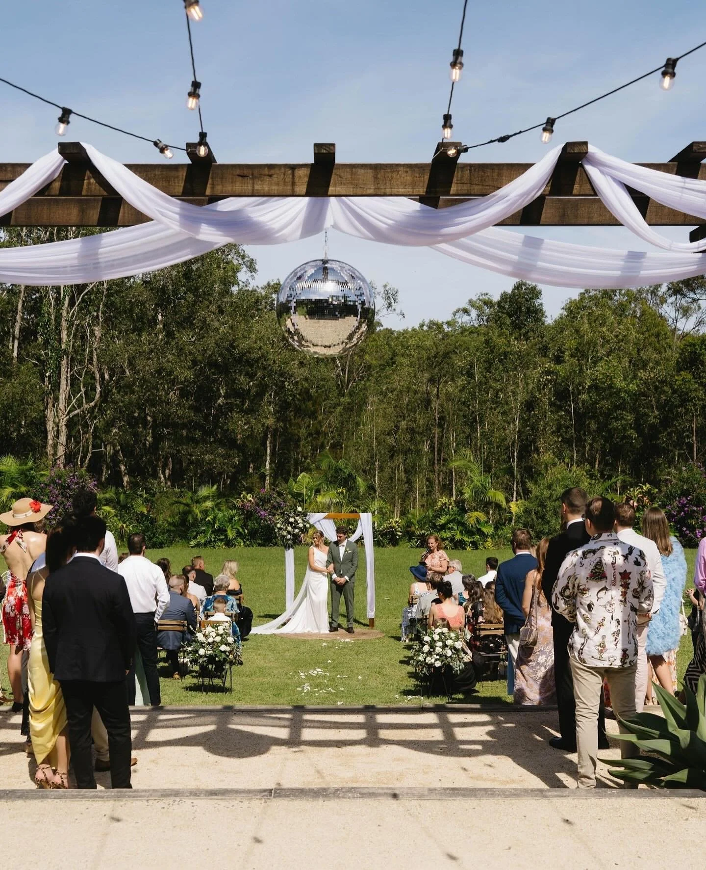 Outdoor wedding ceremony with a bride and groom standing under an arch, surrounded by guests seated on either side, in a lush garden setting with trees and greenery.