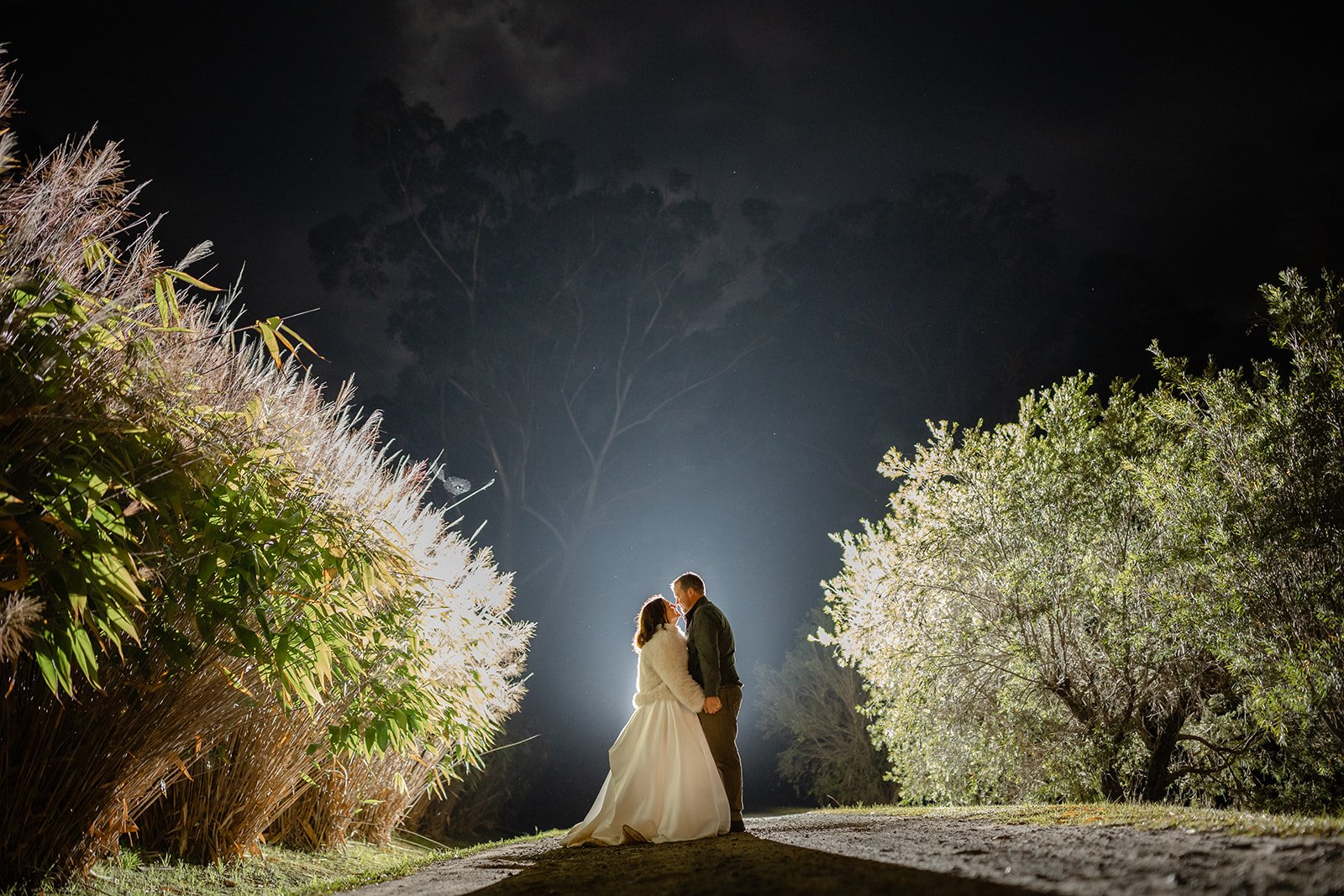A couple on their wedding night stands close, sharing a romantic moment illuminated by a bright light behind them, surrounded by trees and bushes at night.