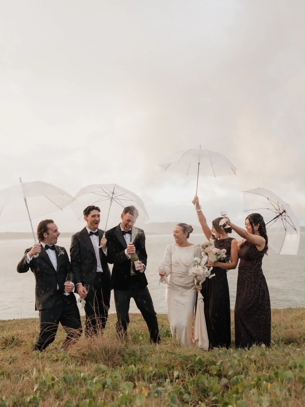 Group of people in wedding attire celebrating outdoors with umbrellas and champagne on a cloudy day.