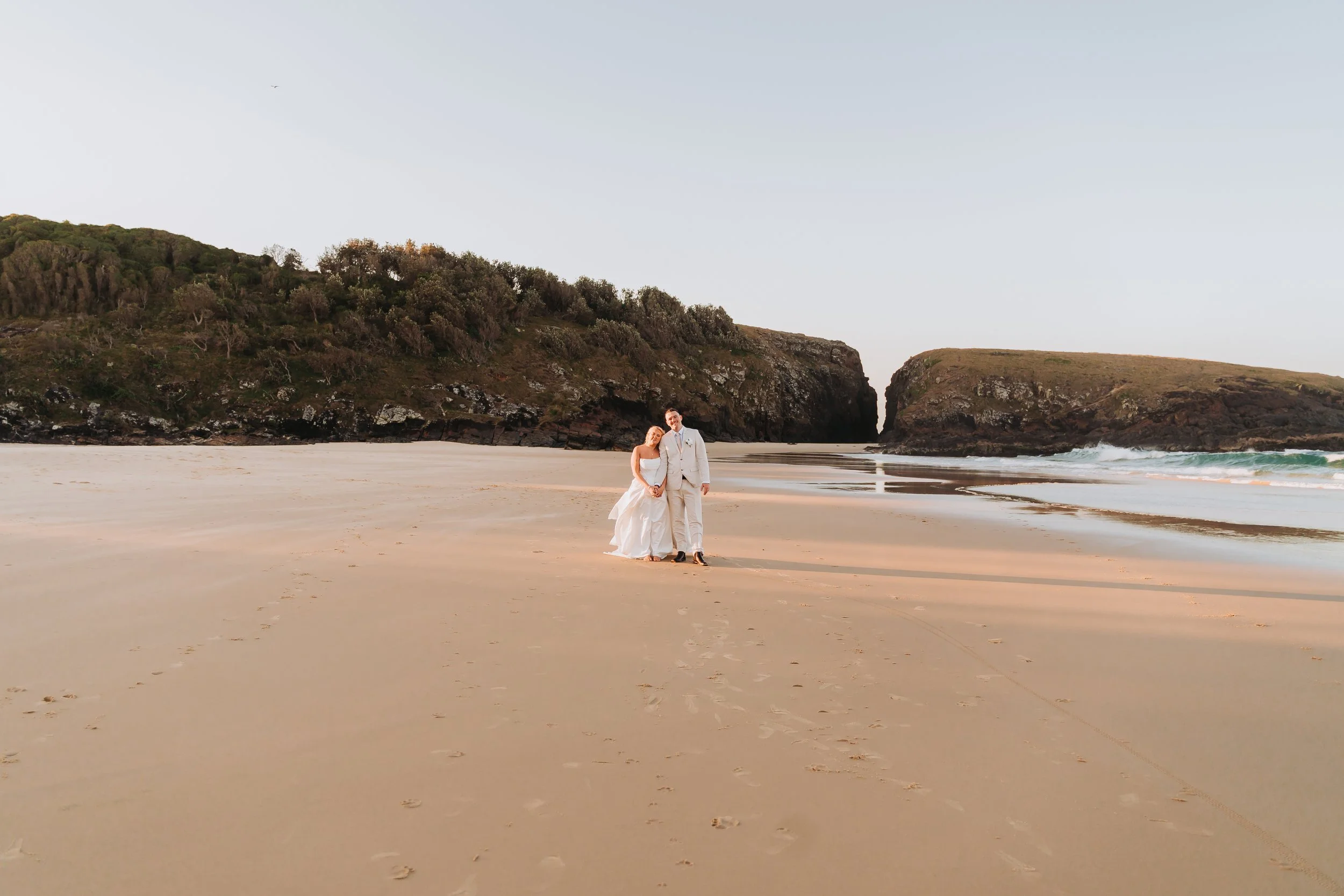 A couple in wedding attire standing hand in hand on a sandy beach with cliffs and greenery in the background.