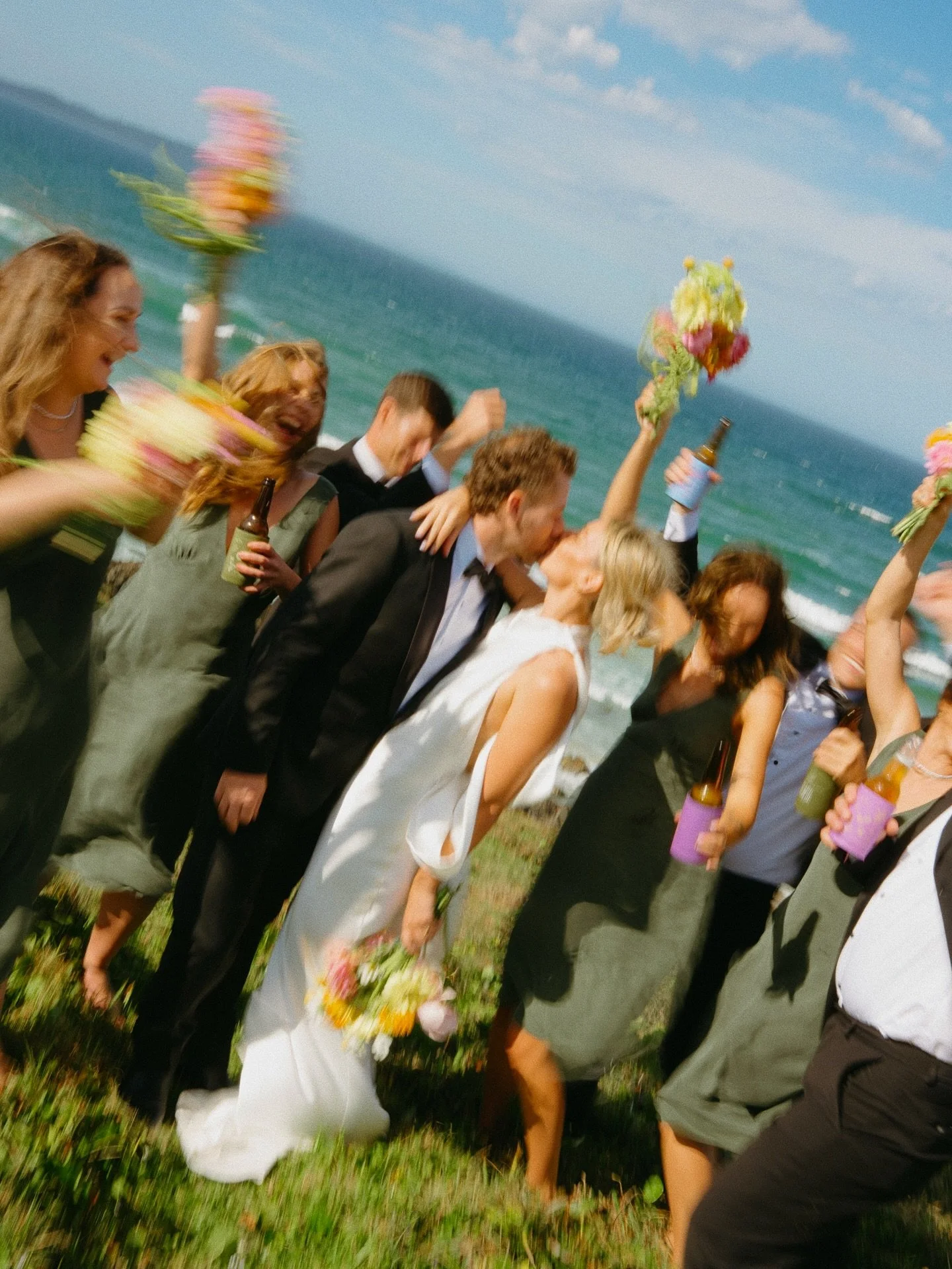 People at a wedding on a beach, celebrating with flower bouquets and drinks, kissing and smiling.