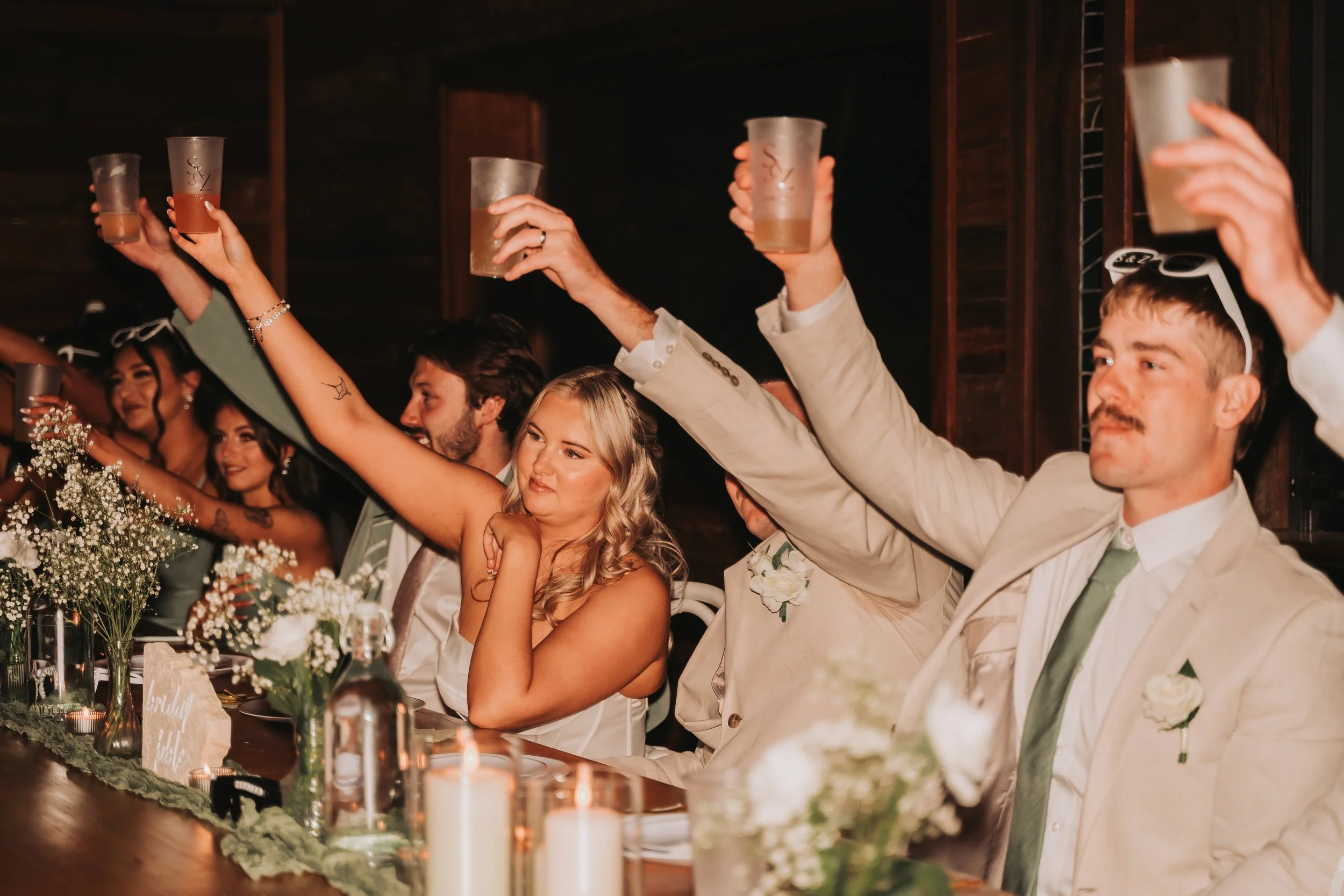 Guests at a wedding celebration raise their glasses in a toast at a long decorated table with flowers and candles.