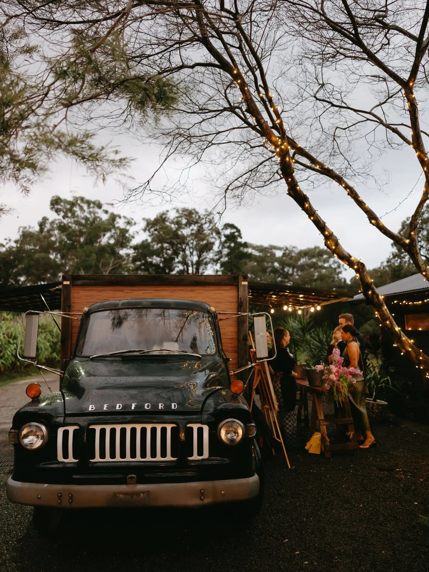 A vintage Bedford truck with a wooden cargo bed is parked outdoors with a group of women gathered around a table with flowers and decorations. Trees with fairy lights are in the background, and it appears to be dusk or early evening.